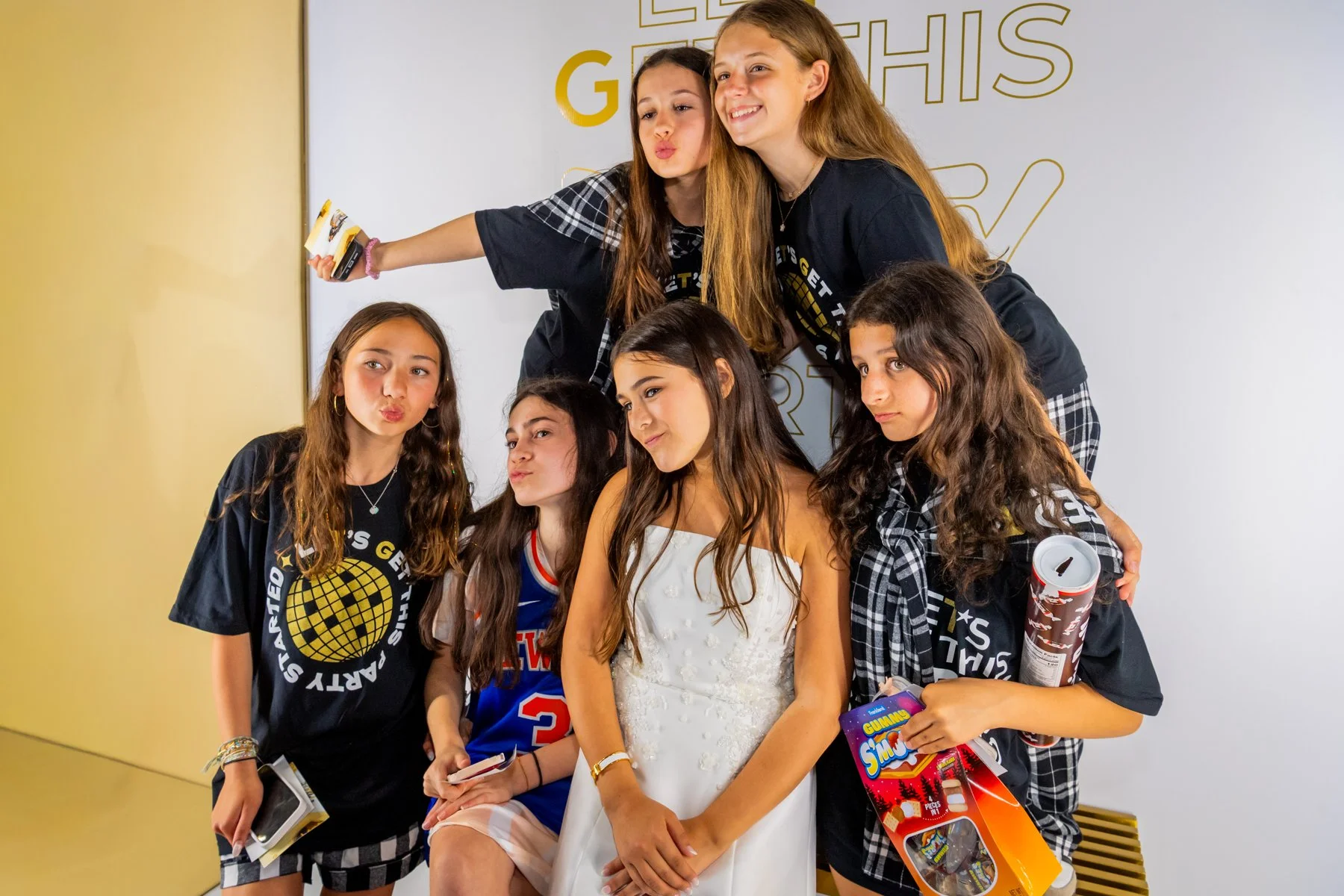 A group of young girls and women posing together for a photo at an event, holding snacks and toys, with some making playful expressions. They are wearing casual clothes, and the background features a white wall with yellow text.