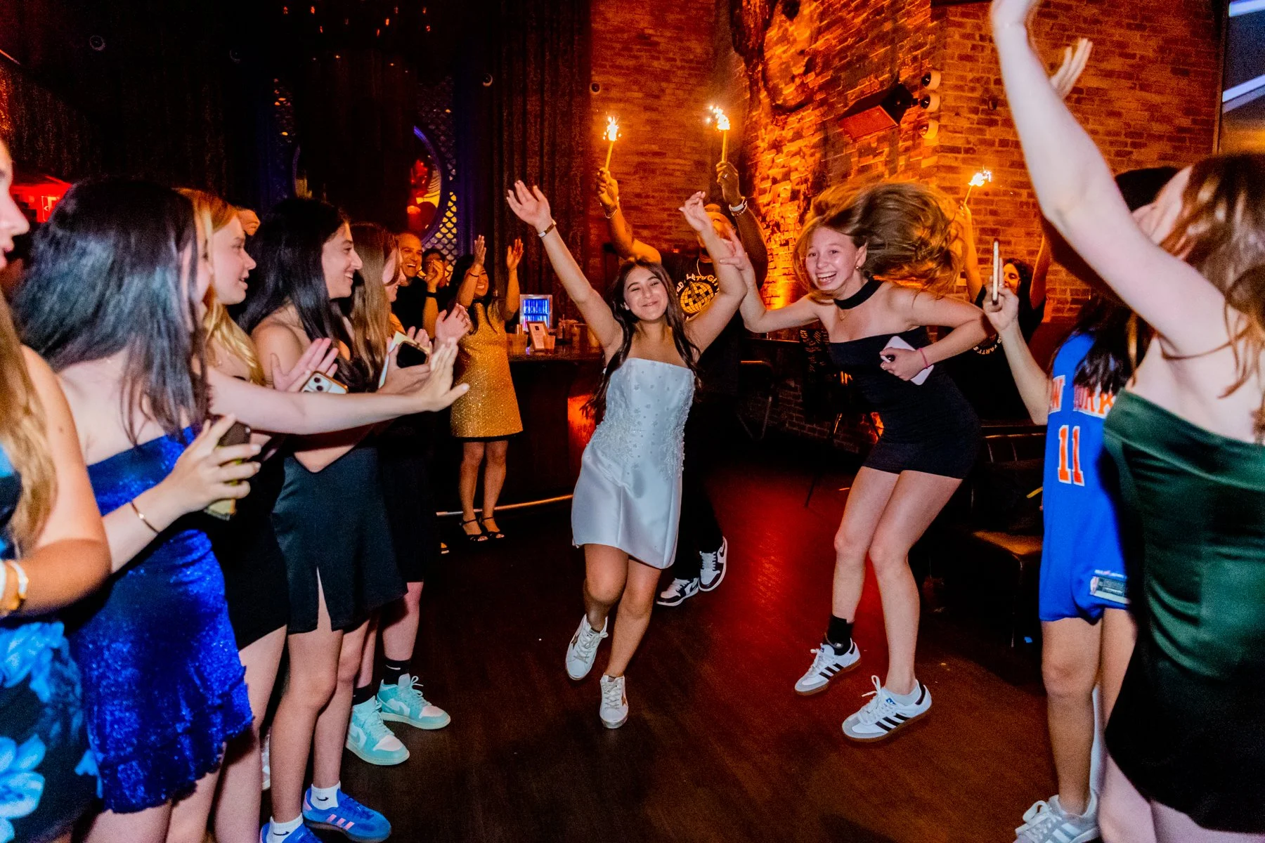 Young women dancing and celebrating at a party, with some holding sparklers, in a dimly lit venue with brick walls.