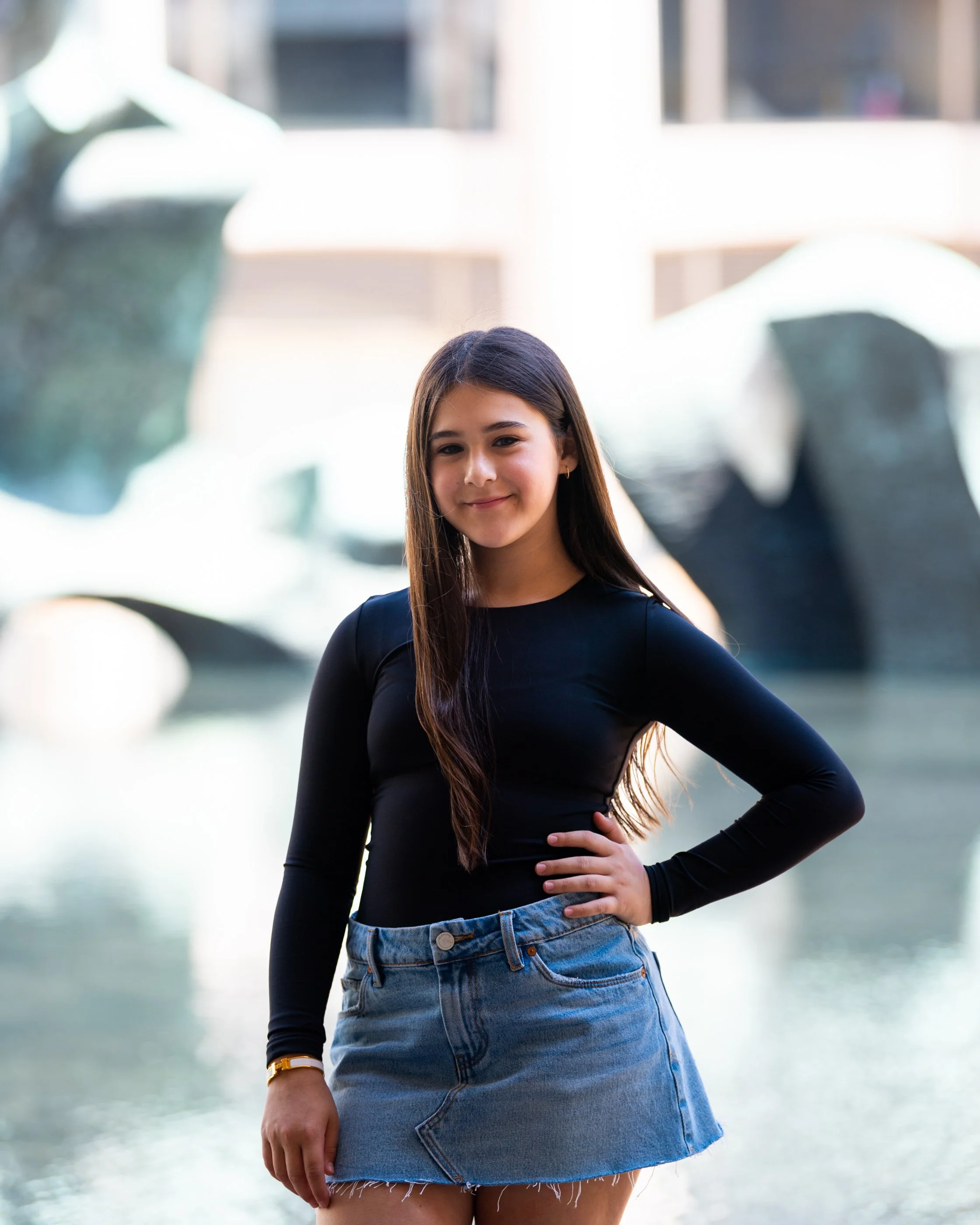 A young woman with long brown hair wearing a black long-sleeve top and a blue denim skirt standing outdoors near water with water fountains in the background.