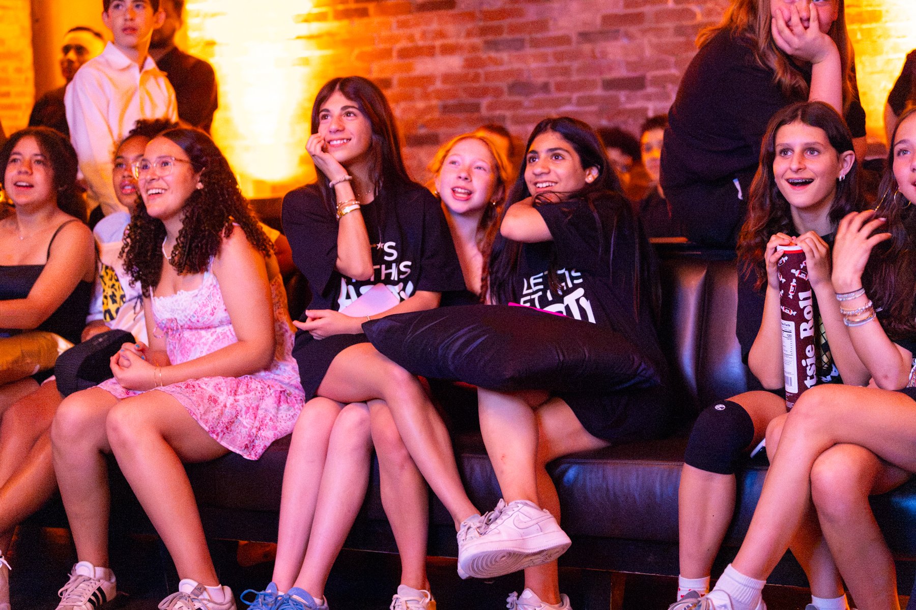 Group of young girls sitting on a black couch, watching something and smiling, in a dimly lit room with exposed brick walls, some holding snacks.