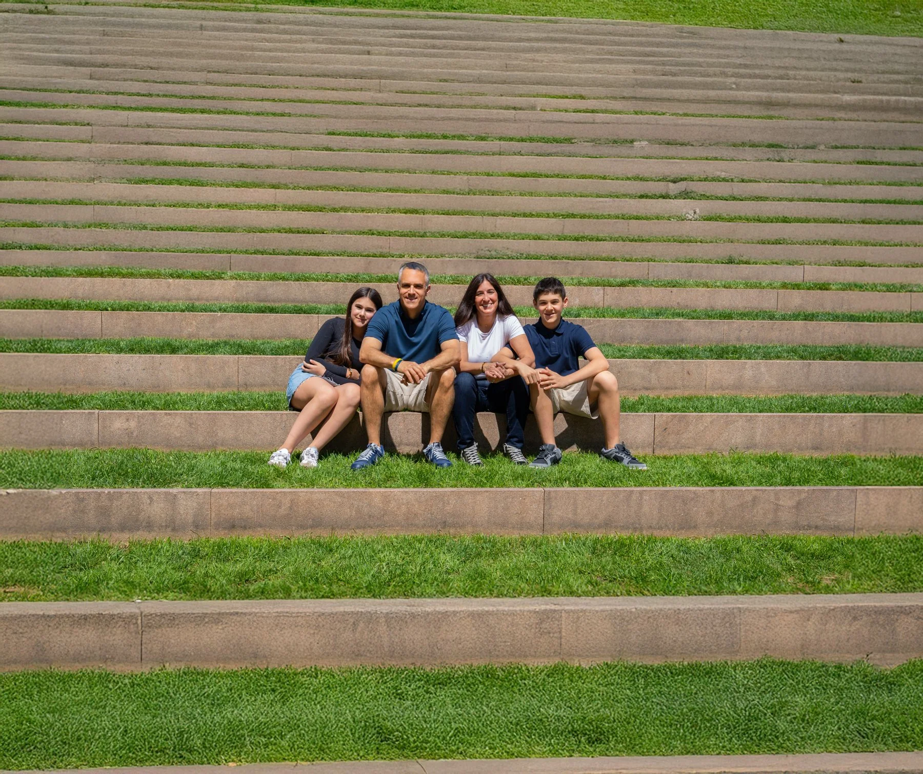 A family of four sitting on stone steps outdoors, smiling at the camera. The family includes a teenage girl, a middle-aged man, a woman, and a teenage boy. The steps are wide with patches of grass growing in between.