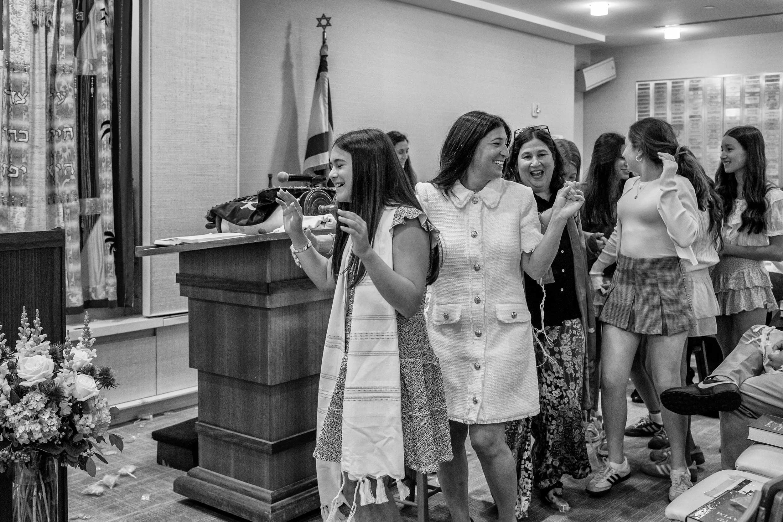 A group of women and girls celebrating at a gathering, some smiling and holding hands, in a room with a Jewish flag and flowers.