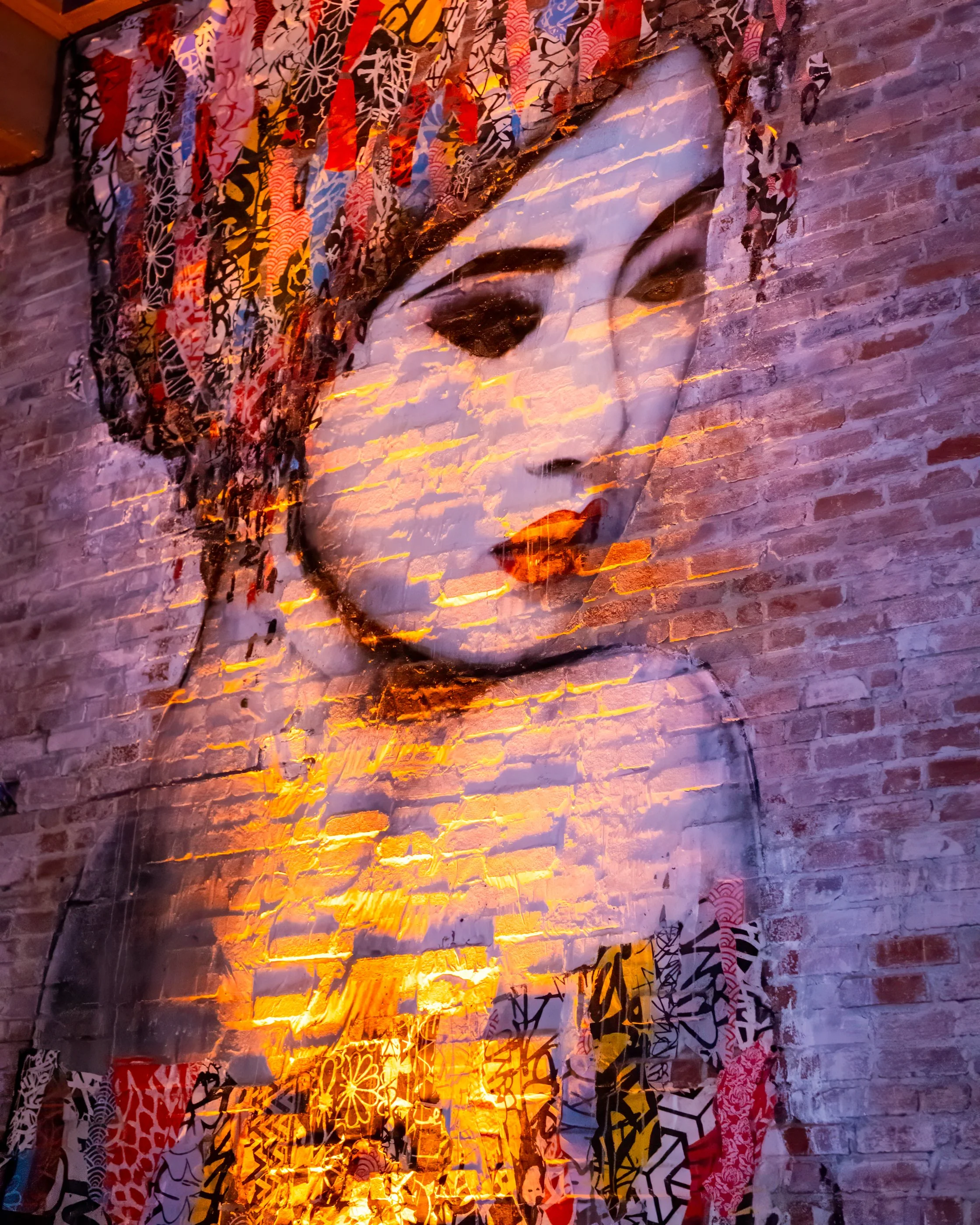 Colorful mural of a woman's face with traditional Japanese hair accessories, mounted on a brick wall, illuminated with orange lighting.