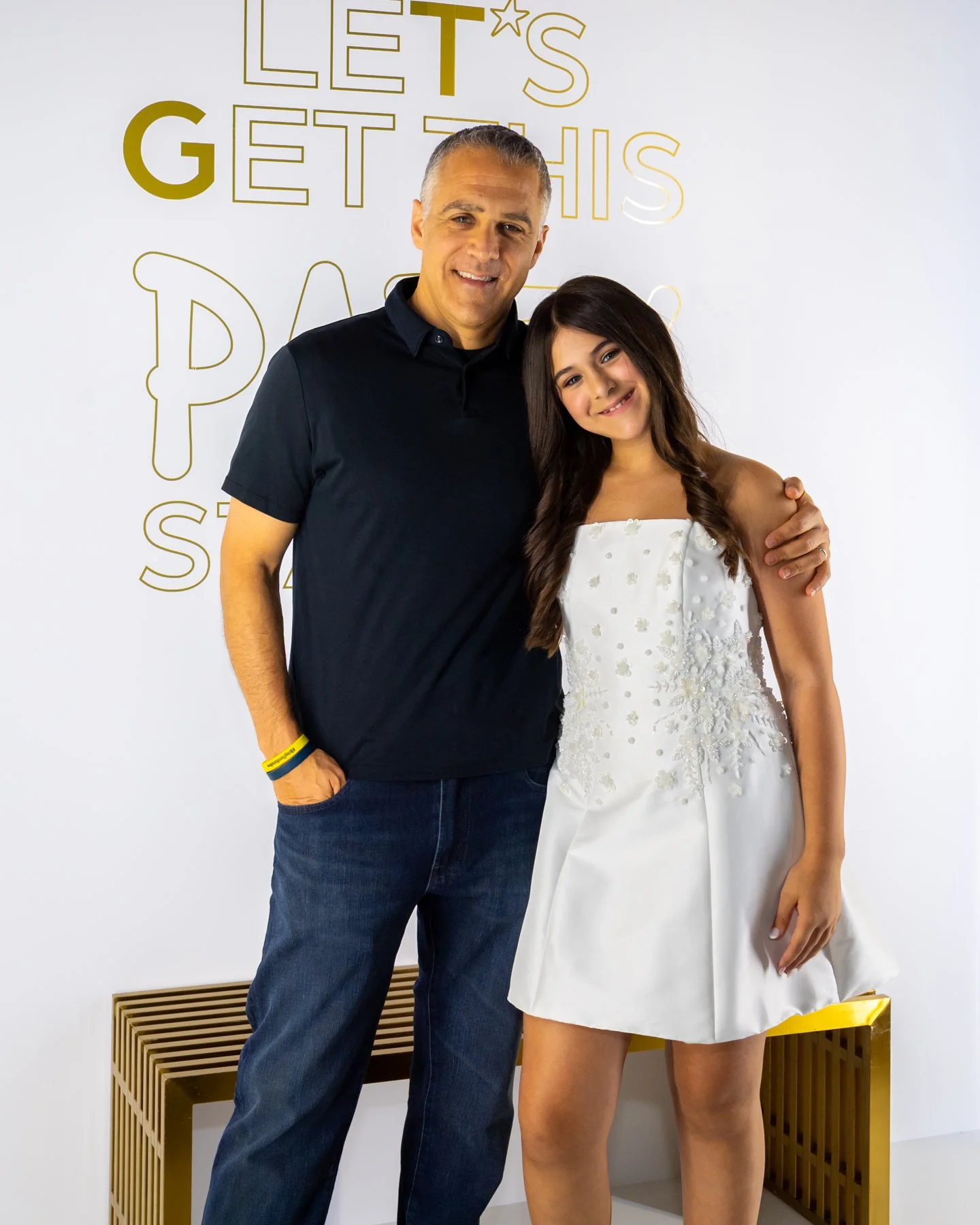 A man and a young woman pose together for a photo in front of a white wall with gold lettering saying "Let's get this day." The man is wearing a black polo shirt and jeans, and the woman is in a white dress with floral embroidery.