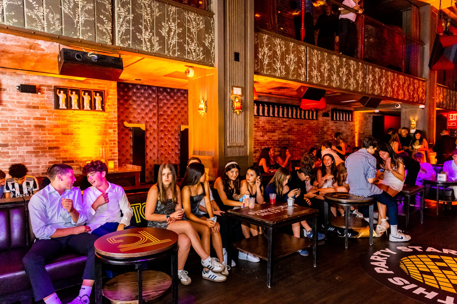 A crowded indoor party scene with young people socializing and sitting around tables in a dimly lit venue with warm lighting, wooden accents, brick walls, and decorative lighting fixtures.
