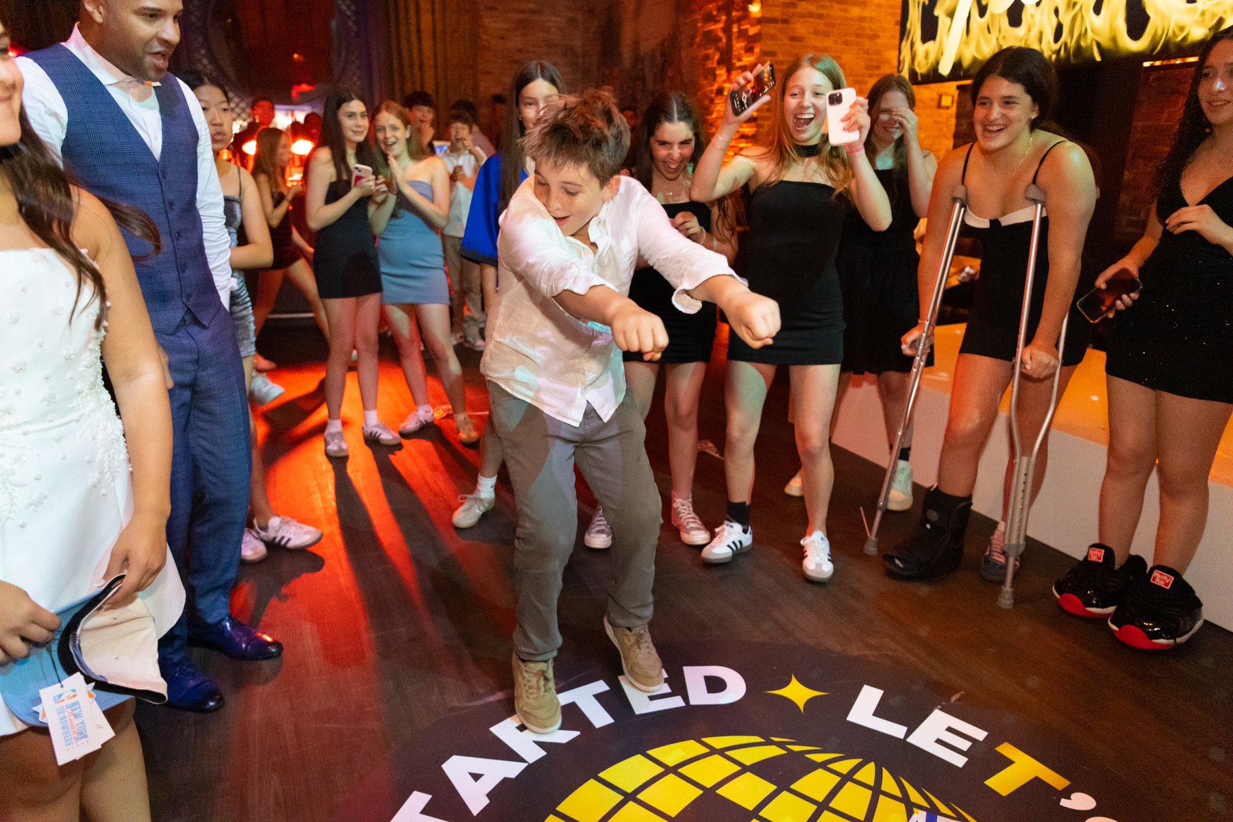A group of young people at a TEDx event, with one person dancing in the center while others watch and cheer. Some people are taking photos or videos. The setting is indoors with a wooden floor and brick walls.