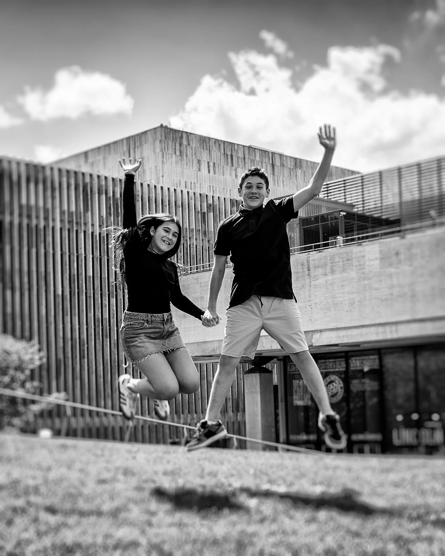 Two kids, a boy and a girl, are jumping over a slackline in an urban outdoor area with modern buildings in the background, captured in black and white.