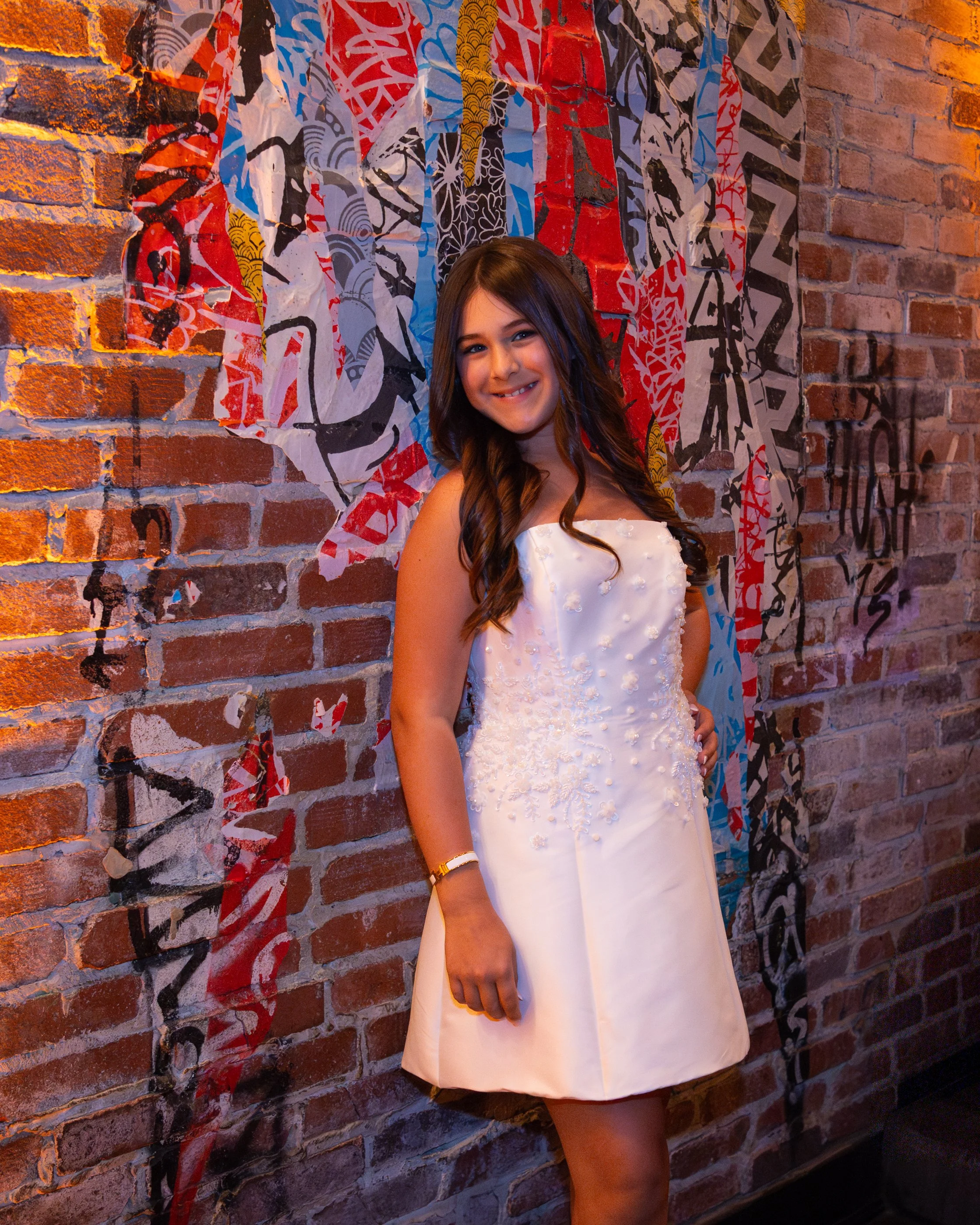 A young girl with long brown hair in a white dress smiling, standing in front of a red brick wall with colorful abstract graffiti art.