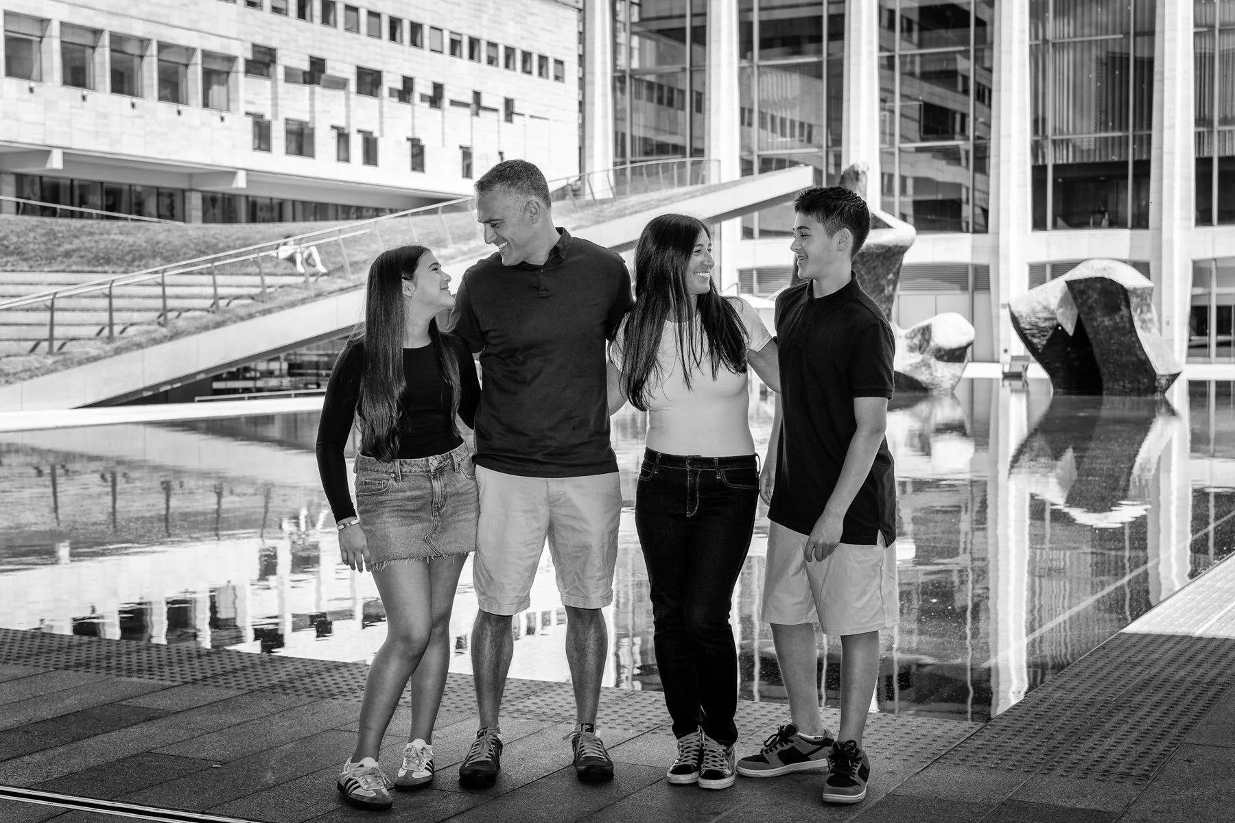 Four family members, two females and two males, stand together by a water feature in an urban plaza, smiling and talking with each other. Modern buildings are in the background.