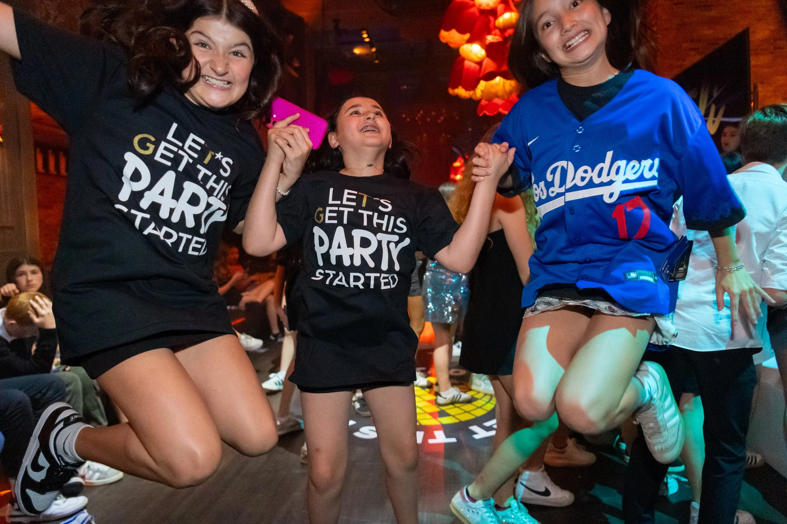 Three young girls at a lively party, smiling and dancing with their arms joined. They are wearing casual clothes including black shorts and sneakers, and two have black t-shirts with 'Let's Get This Party Started' printed on them. The scene is indoor