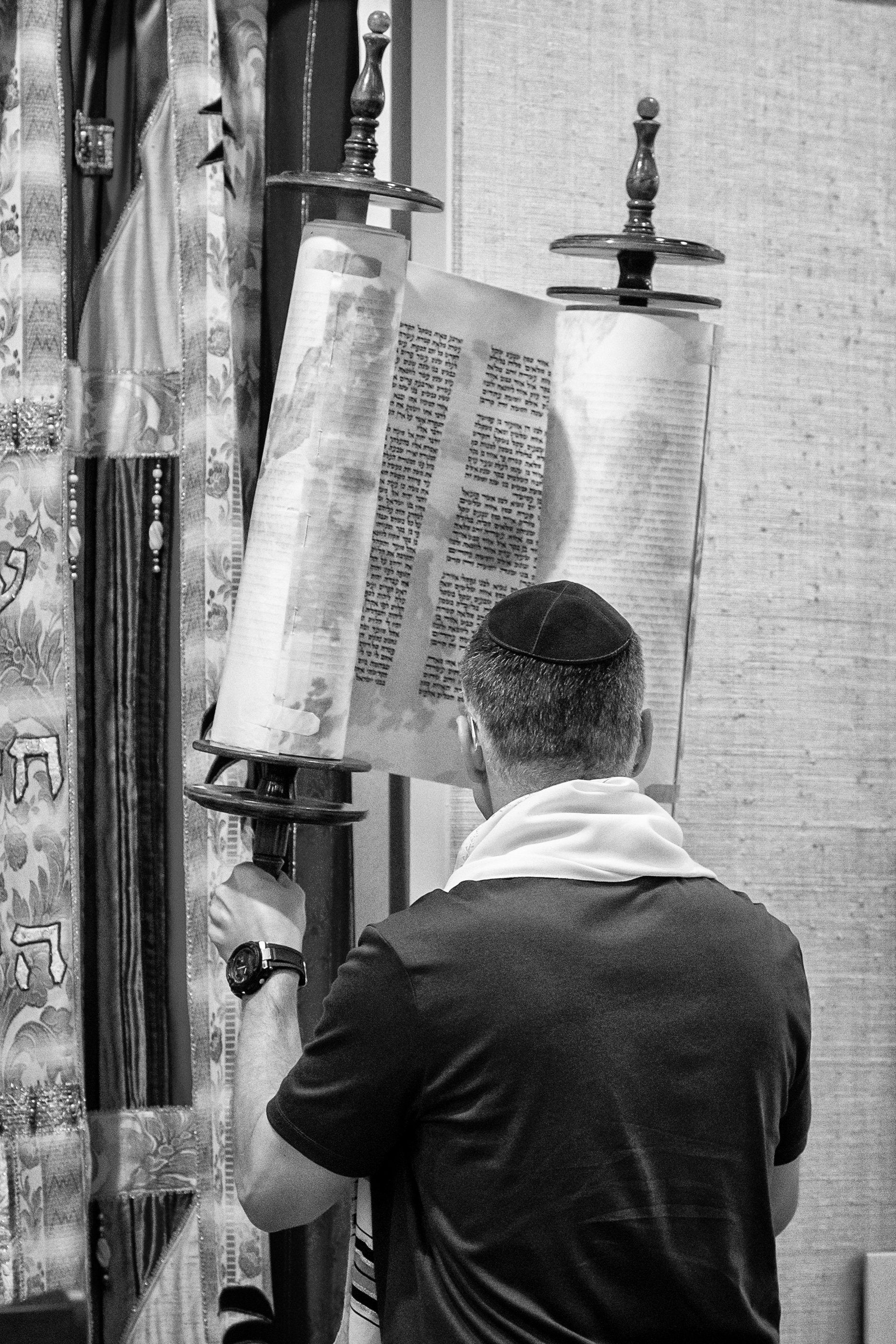 A man wearing a kippah and a watch is standing in front of a mirror, holding a prayer book during prayer. The room has decorative curtains and textured wall panels.