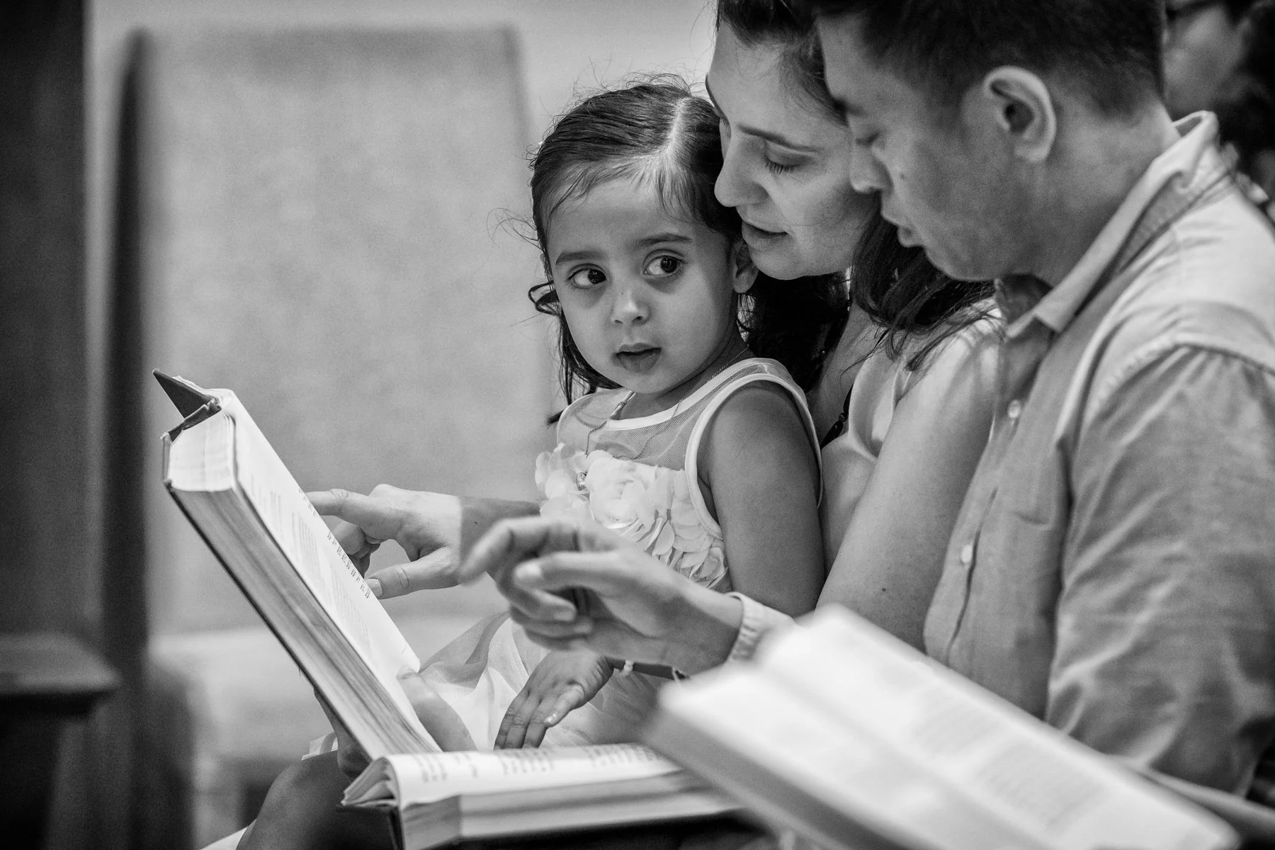 A young girl sitting between two adults, looking at a book, in a grayscale photograph.