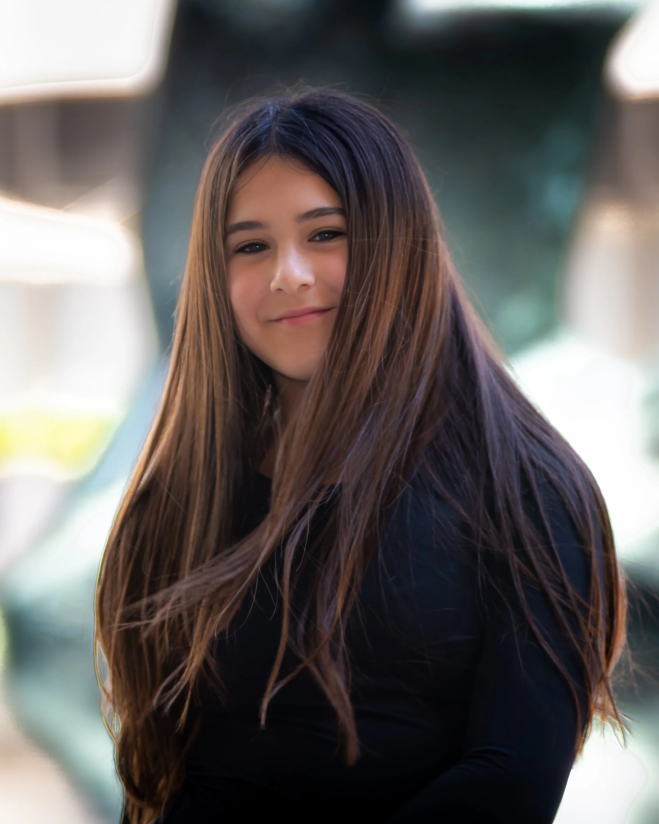 A young woman with long, straight brown hair. She is smiling softly and looking slightly to the side. The background is blurred with bright light and hints of greenery.