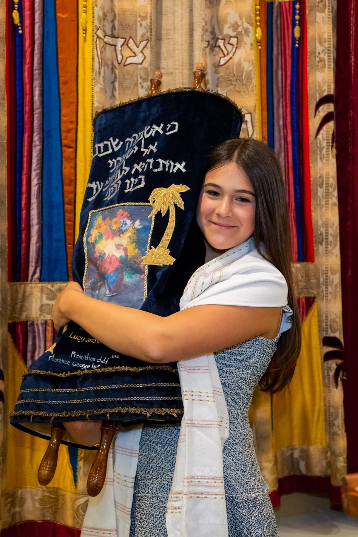 A young girl with long brown hair smiling and hugging a Torah scroll in a synagogue with colorful curtains in the background.