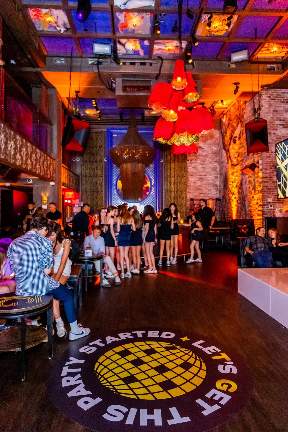 A lively nightclub scene with groups of young people socializing and dancing. The space features colorful lighting, a brick wall, and a prominent chandelier. A large circular logo on the floor reads 'Party Started Let's' with a globe graphic.