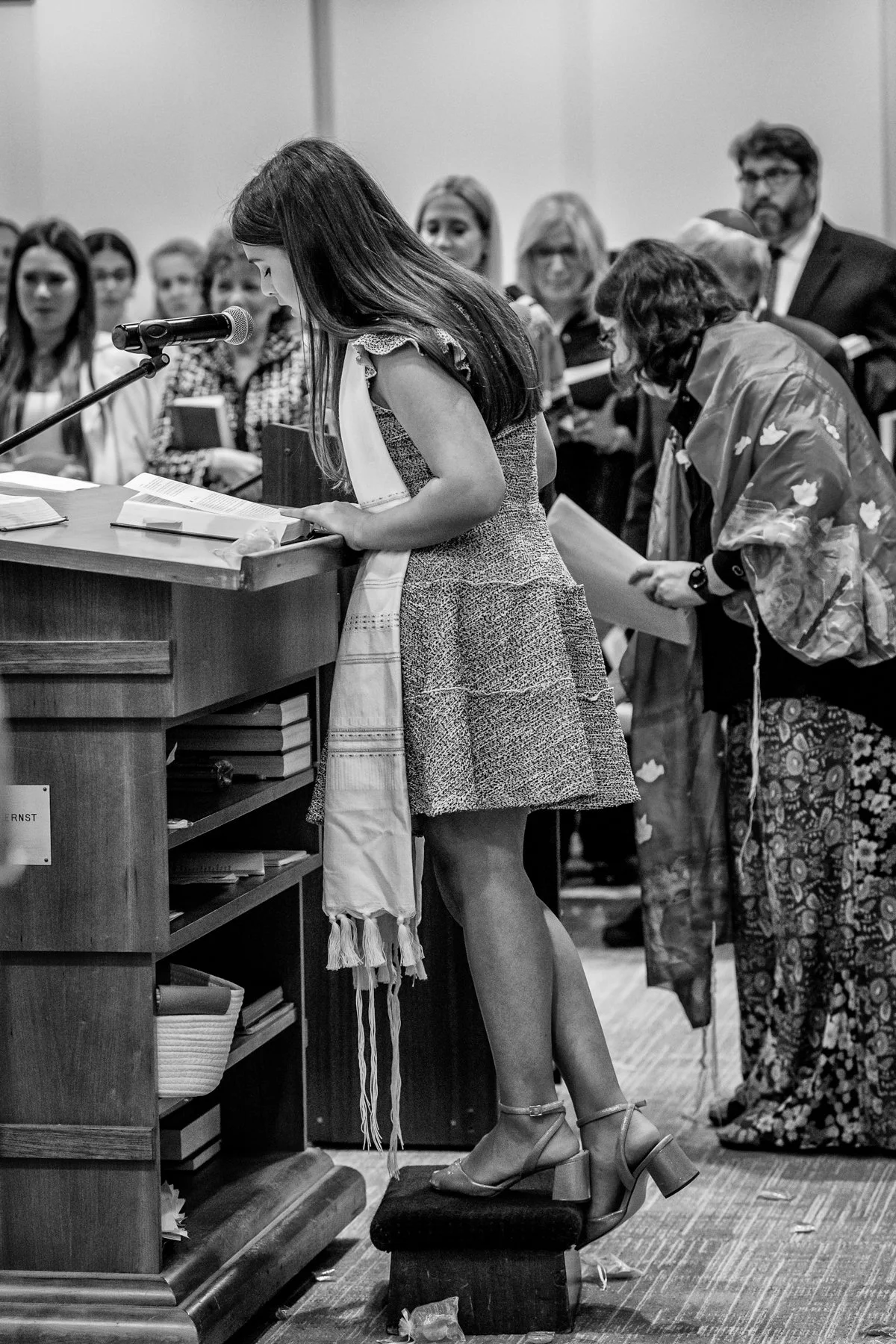 A woman in a dress and heels stands on a footstool reading from a book at a podium, with a microphone, at a gathering with many people listening.