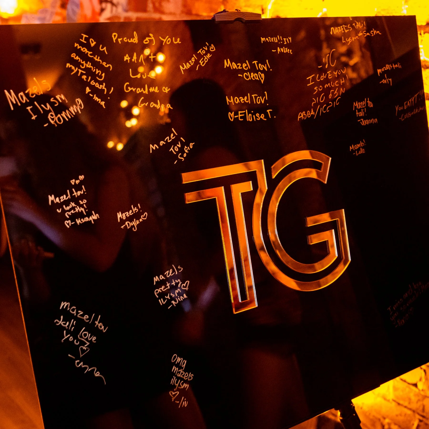 A black object, possibly a photo album or a display board, with numerous handwritten notes and messages in white and orange ink. The background is lit with warm lighting, and some reflections or lights are visible on the surface. The notes contain ex