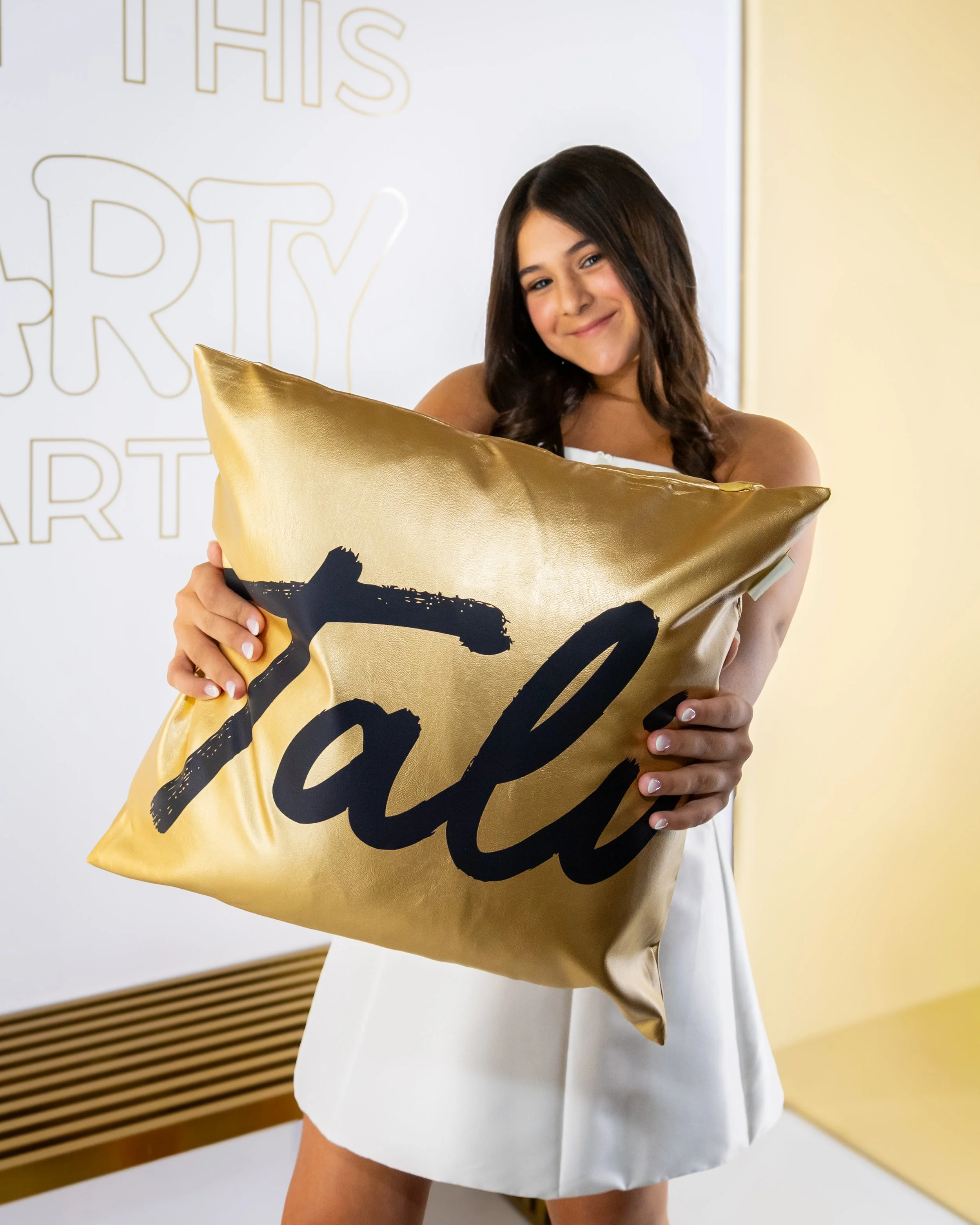A young woman with brown hair smiling and holding a gold-colored pillow with black writing, standing in front of a backdrop with text that reads 'This is my art'.