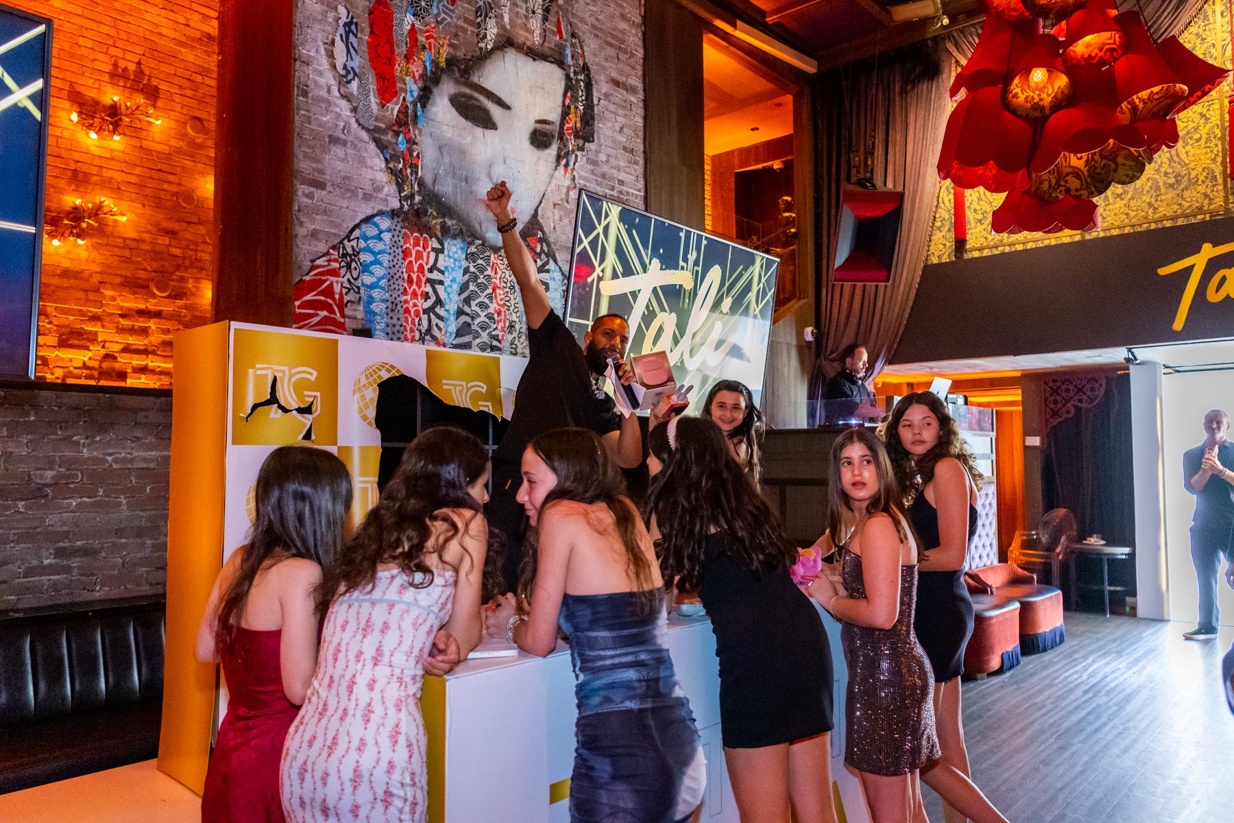 A group of young girls in party dresses gathered around a table at a lively event. A man stands behind the table speaking into a microphone, raising his fist in the air. The background features a large mural of a woman with traditional Asian makeup a