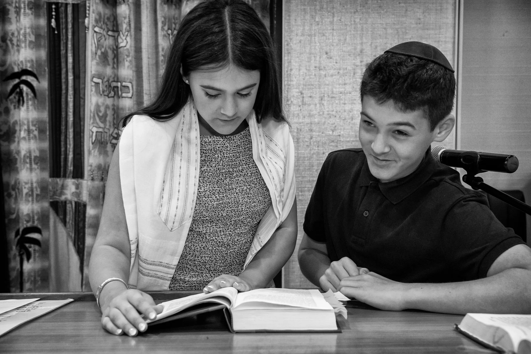 A woman and a boy looking at open books on a table, with a microphone nearby, in an indoor setting.