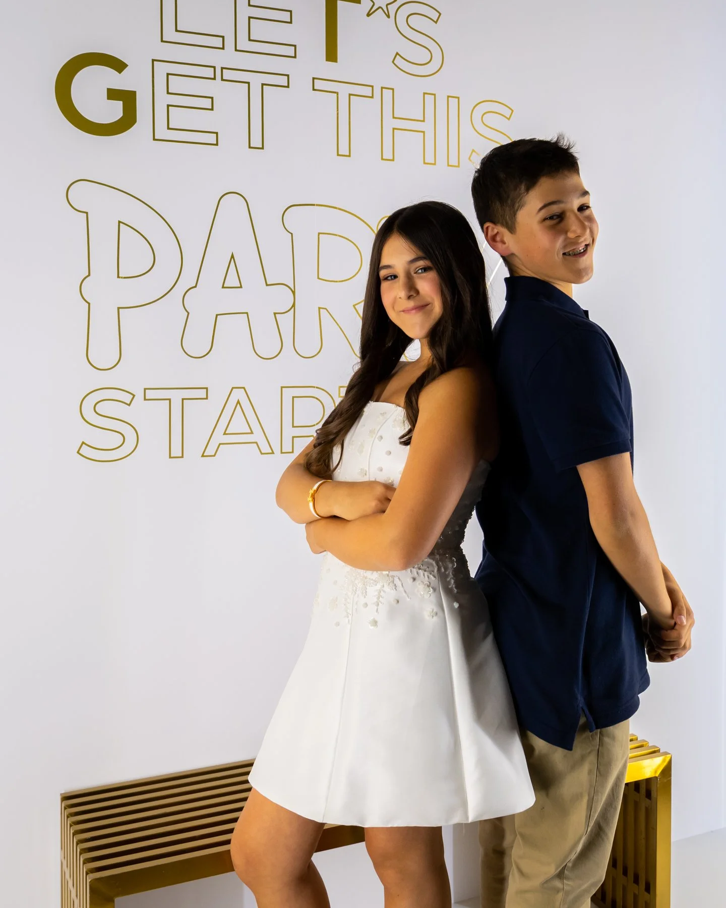 A girl in a white dress and a boy in a navy shirt and khaki pants standing back-to-back in front of a white wall with gold lettering that says 'Let's Get This Party Started.' The girl has long dark hair and is smiling at the camera; the boy has short