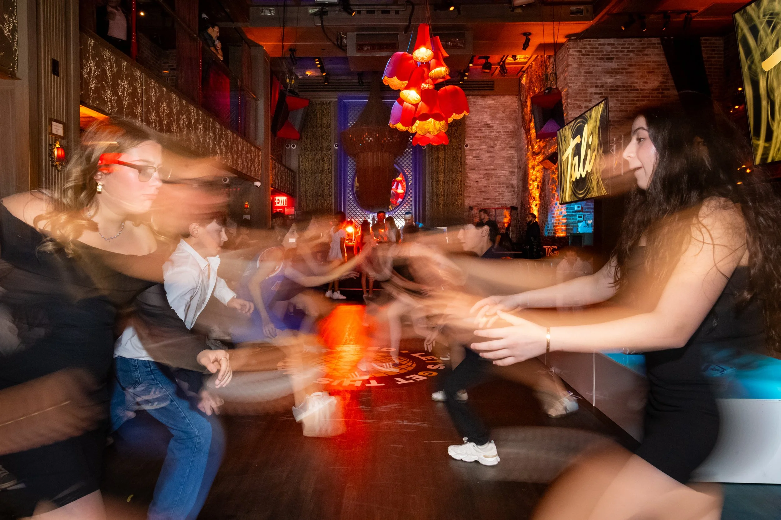 People dancing in a dimly lit nightclub with red lighting, exposed brick walls, and large TV screens.