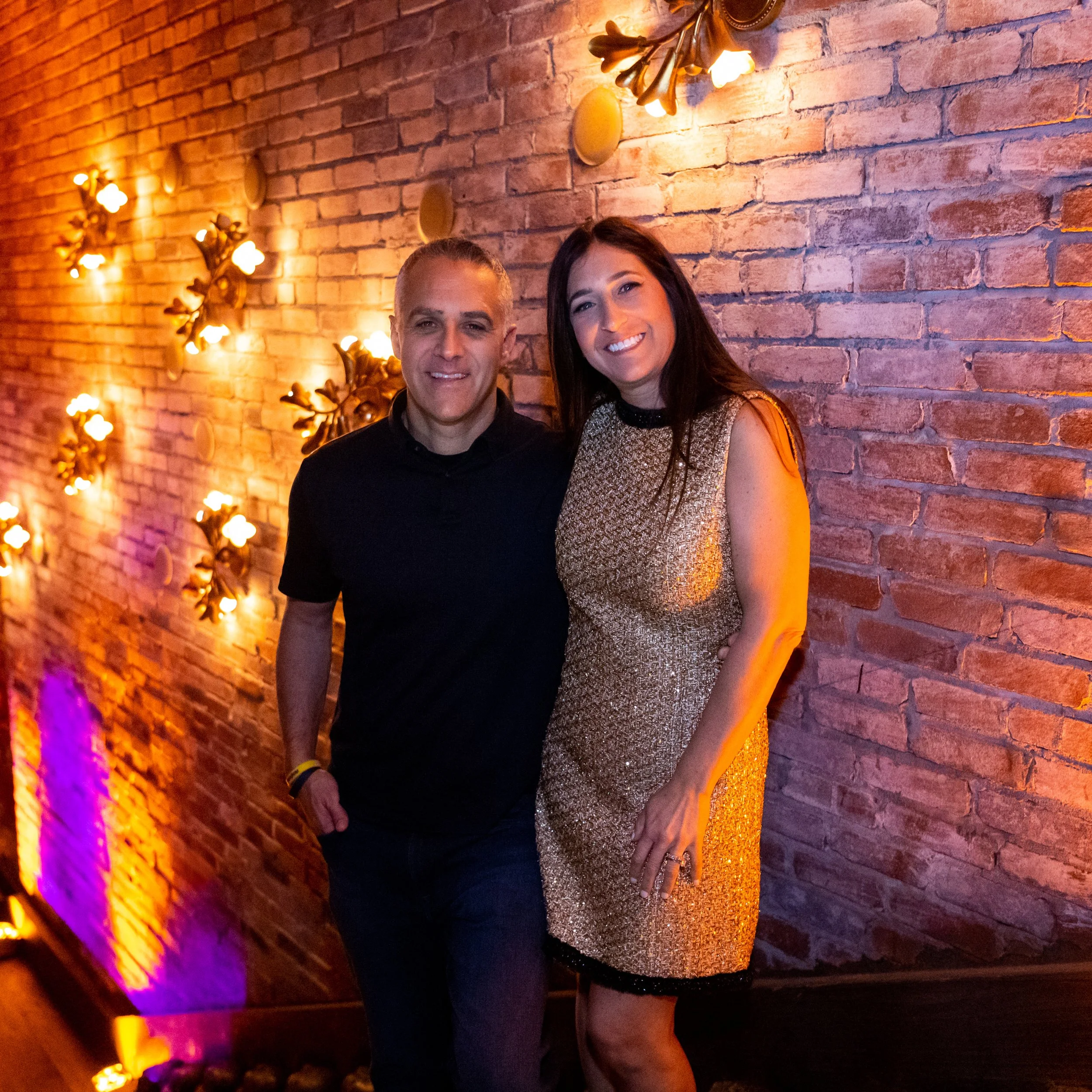 A man and a woman smiling and posing for a photo in front of a brick wall decorated with warm-colored floral wall sconces and purple lighting.