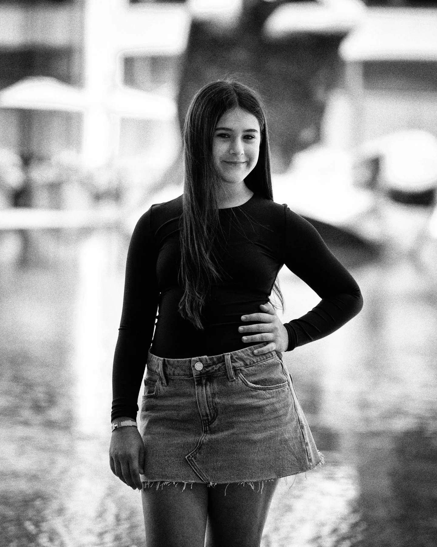 Black and white photo of a young woman standing outdoors in Lincoln Center, smiling with one hand on her hip, wearing a long-sleeve top and denim skirt.