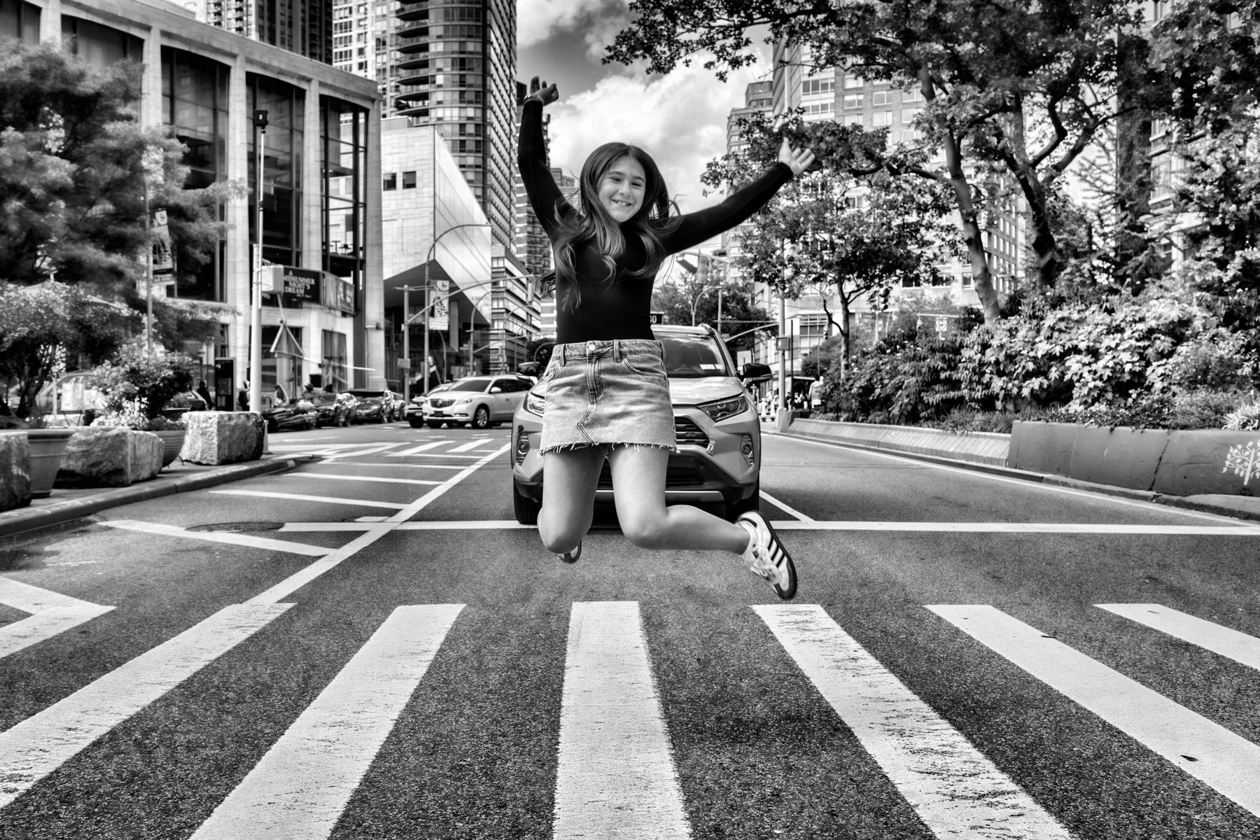 A young woman jumps joyfully in the street of a city, crossing a pedestrian crosswalk with cars and tall buildings in the background. Trees line the sidewalk.