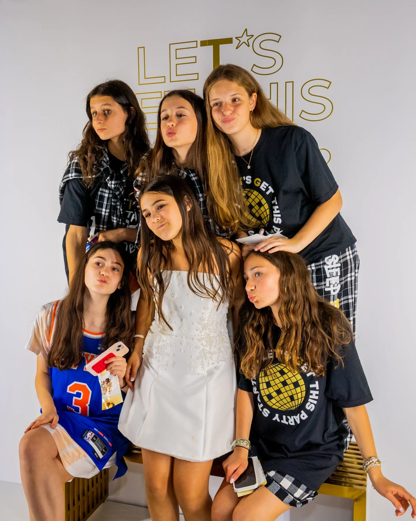 Group of six young girls posing together against a white background with the words 'Let's Get This Party Started' written on it. They are making playful facial expressions, some standing and others sitting, wearing casual and sporty outfits, with one