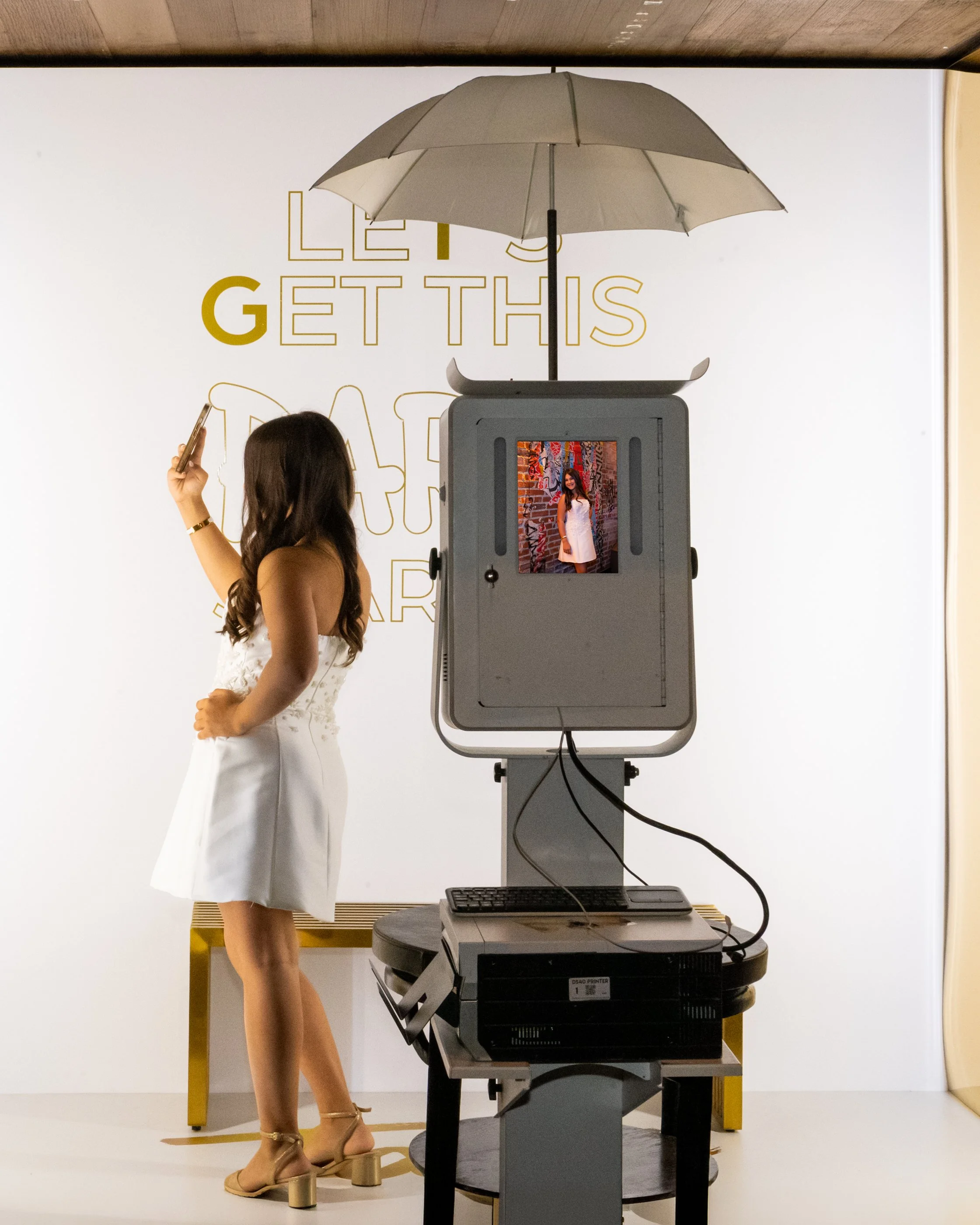 A woman in a white dress taking a photo of herself with a mobile phone in a photo booth. The photo booth has a screen showing her picture, an umbrella overhead, and a large printer below. The background features a white wall with yellow text and desi