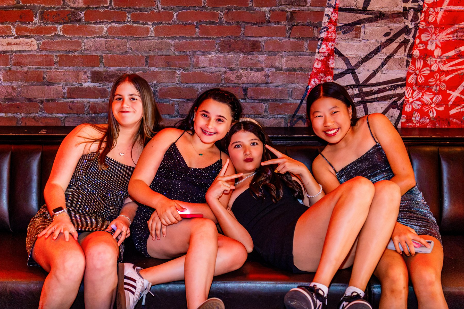 Four young women sitting together on a black leather bench against a brick wall background and a painted floral mural, smiling and posing for a photo, wearing party dresses.