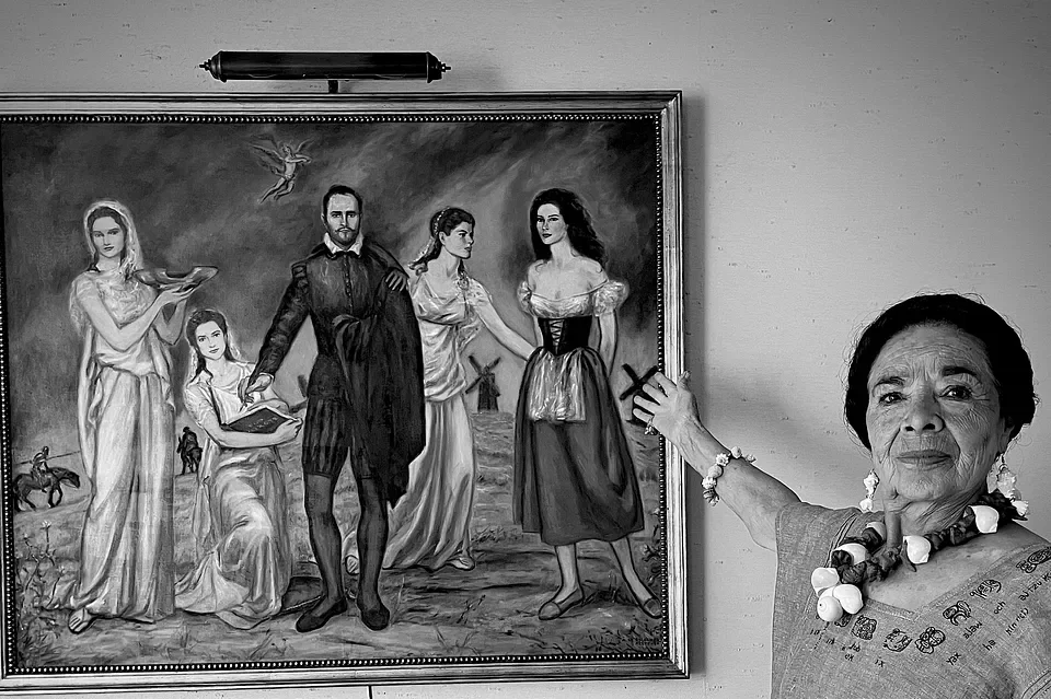 Anciana señalando una pintura en blanco y negro que muestra varias figuras históricas con un paisaje de fondo.