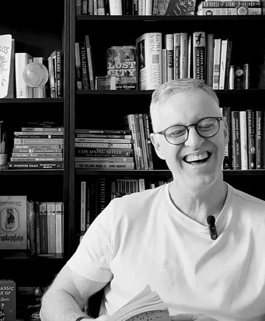 Hombre rubio con gafas, sonriendo y leyendo un libro, con estantería llena de libros en el fondo.