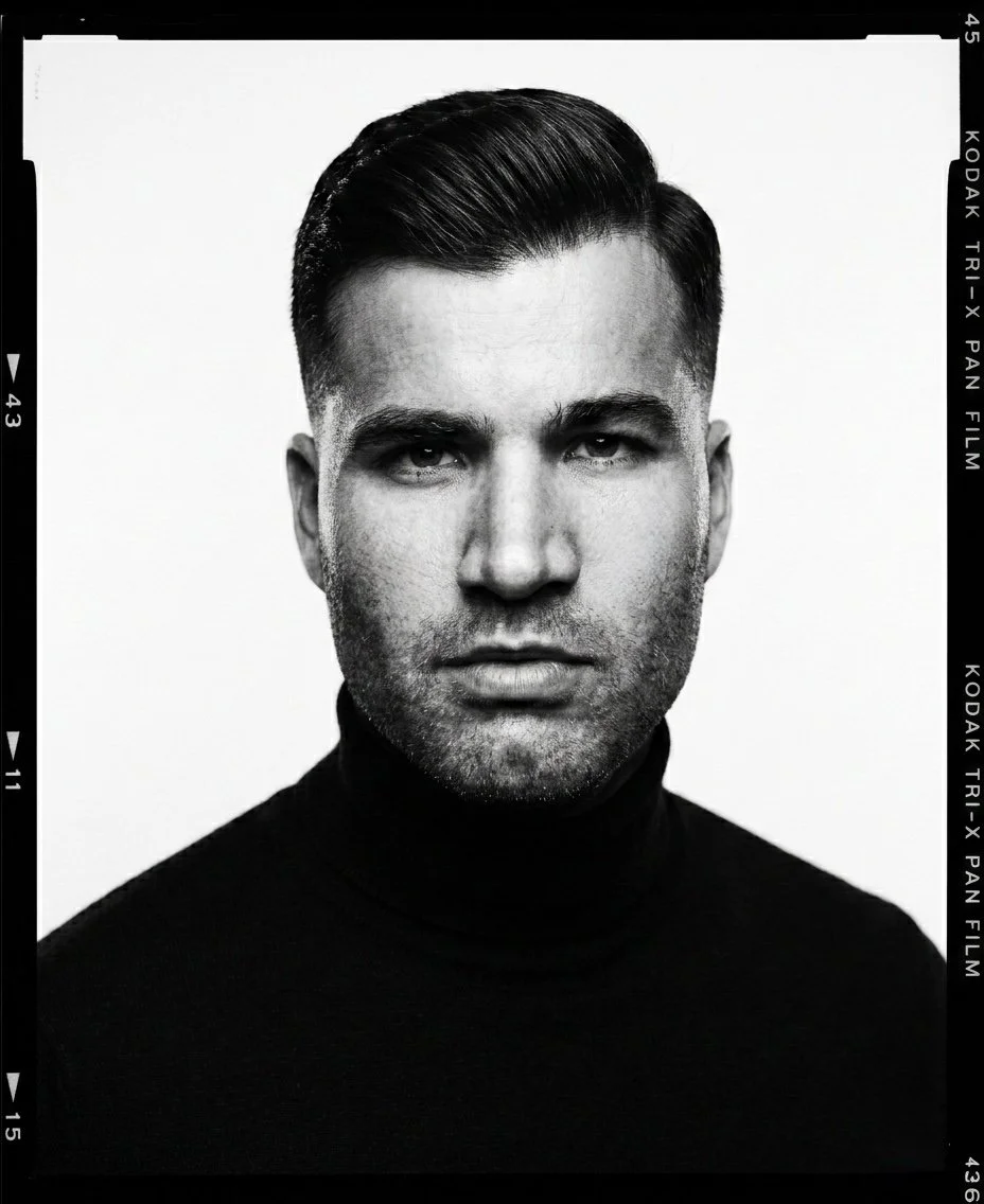 Retrato en blanco y negro de un hombre con cabello oscuro y peinado hacia un lado, usando un suéter negro de cuello alto.
