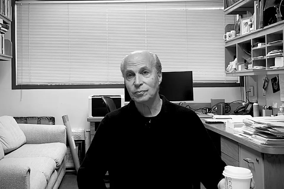 Hombre mayor sentado en una oficina con estantes llenos de papeles y objetos, sosteniendo una taza de café.