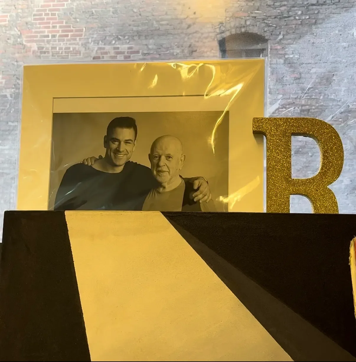 Fotografía de dos hombres, uno joven y otro mayor, sonriendo y abrazados, con decoraciones doradas y una letra 'B' en el fondo, en un ambiente de celebración.