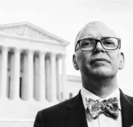 Retrato de un hombre con barba y gafas, vestido con esmoquin y pajarita, frente a un edificio gubernamental clásico.