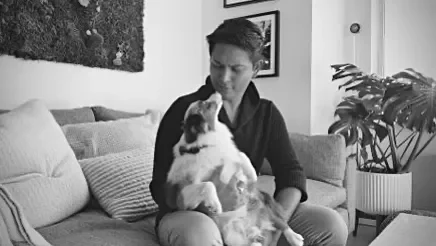 Una mujer sentada en un sofá, sosteniendo y acariciando a un perro grande con manchas negras y blancas.