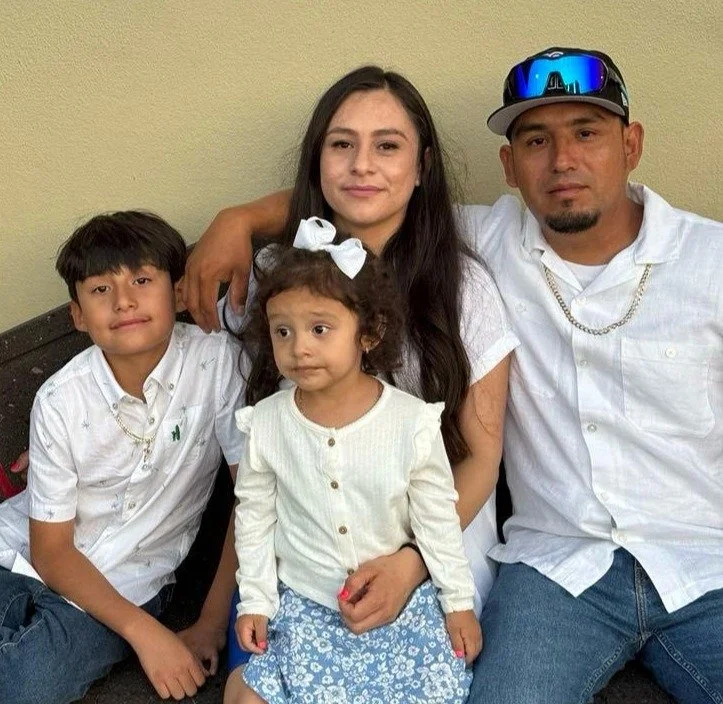 A family of four sitting together, the mother and father with two young children, in front of a yellow wall.