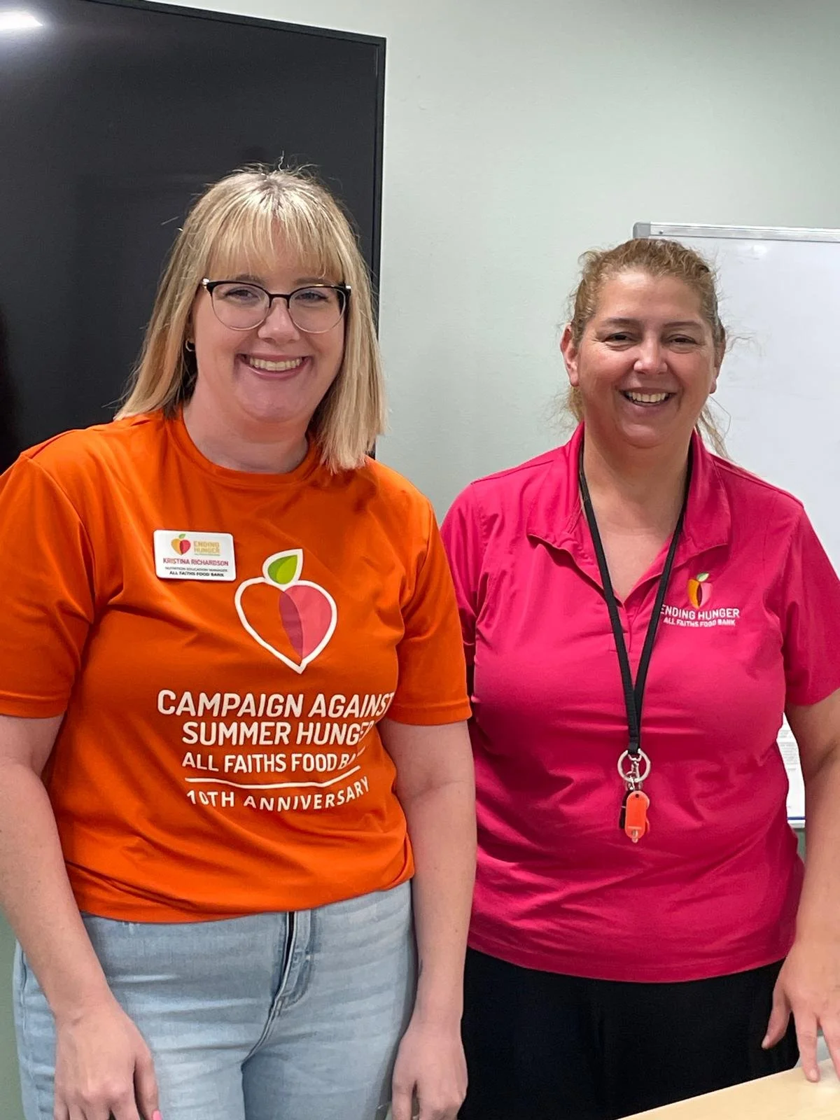 Two women standing side by side, smiling. The woman on the left has blonde hair and wears glasses, an orange T-shirt with a campaign logo and text, and light jeans. The woman on the right has curly hair tied back, wears a pink collared shirt with a logo, and a lanyard around her neck. They are in an indoor setting with a gray wall and a whiteboard behind them.