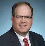 A professional man wearing glasses, a navy blazer, white shirt, and red striped tie, smiling against a plain background.