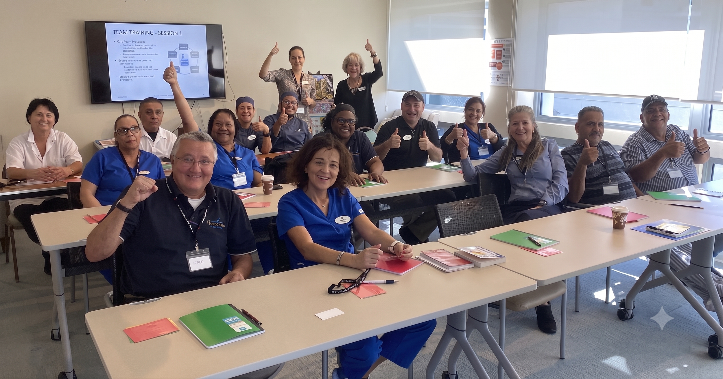 A group of diverse people attending a team training session in a conference room. They are smiling, giving thumbs up, and sitting at tables with books, notebooks, and pens. A presentation slide on a screen reads 'TEAM TRAINING - SESSION 1,' with bullet points about care team protocols. The room has large windows letting in natural light.