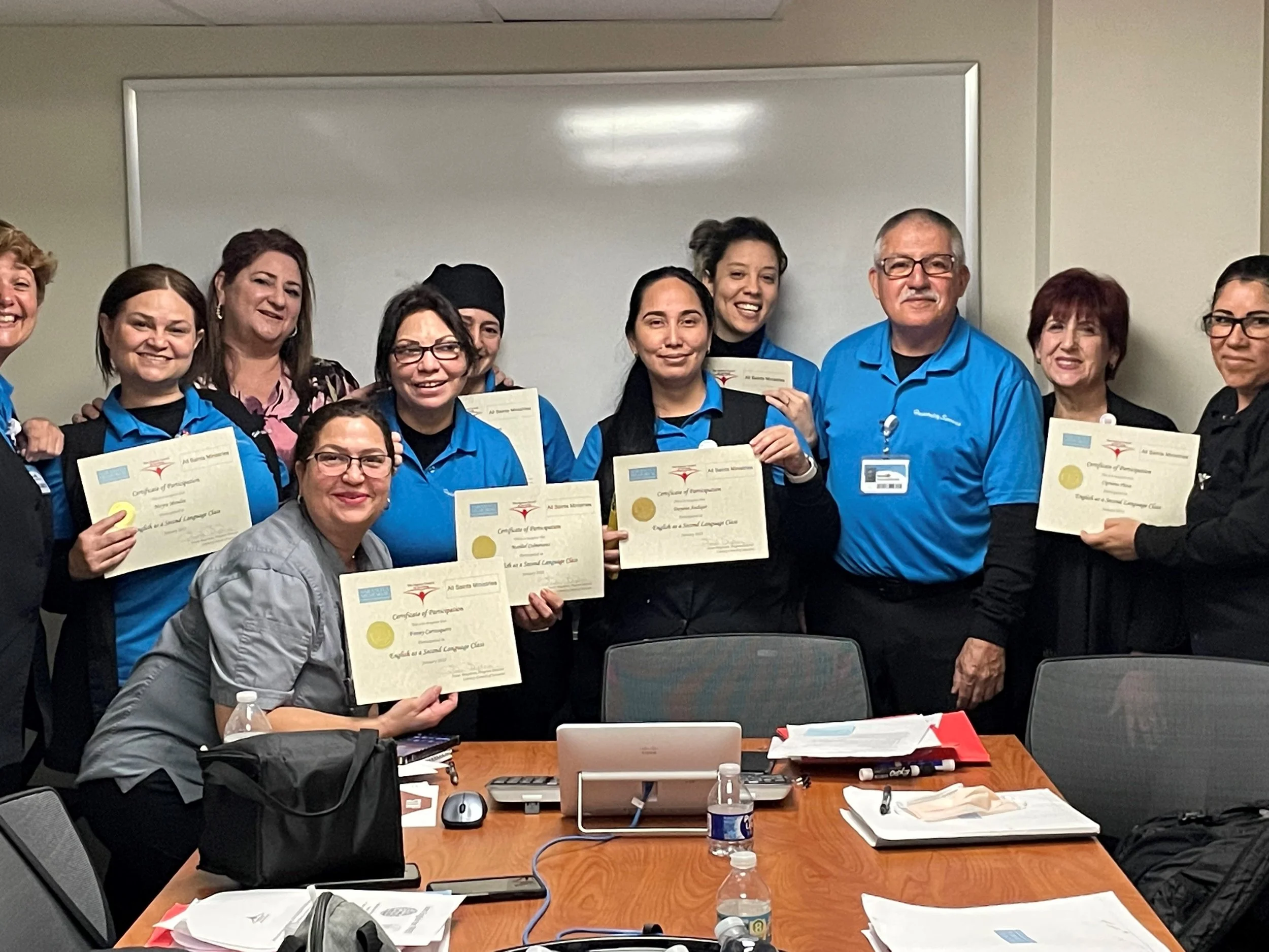 Group of diverse people, mainly women, smiling and holding certificates in a classroom or conference room setting, with a table, papers, and a whiteboard in the background.