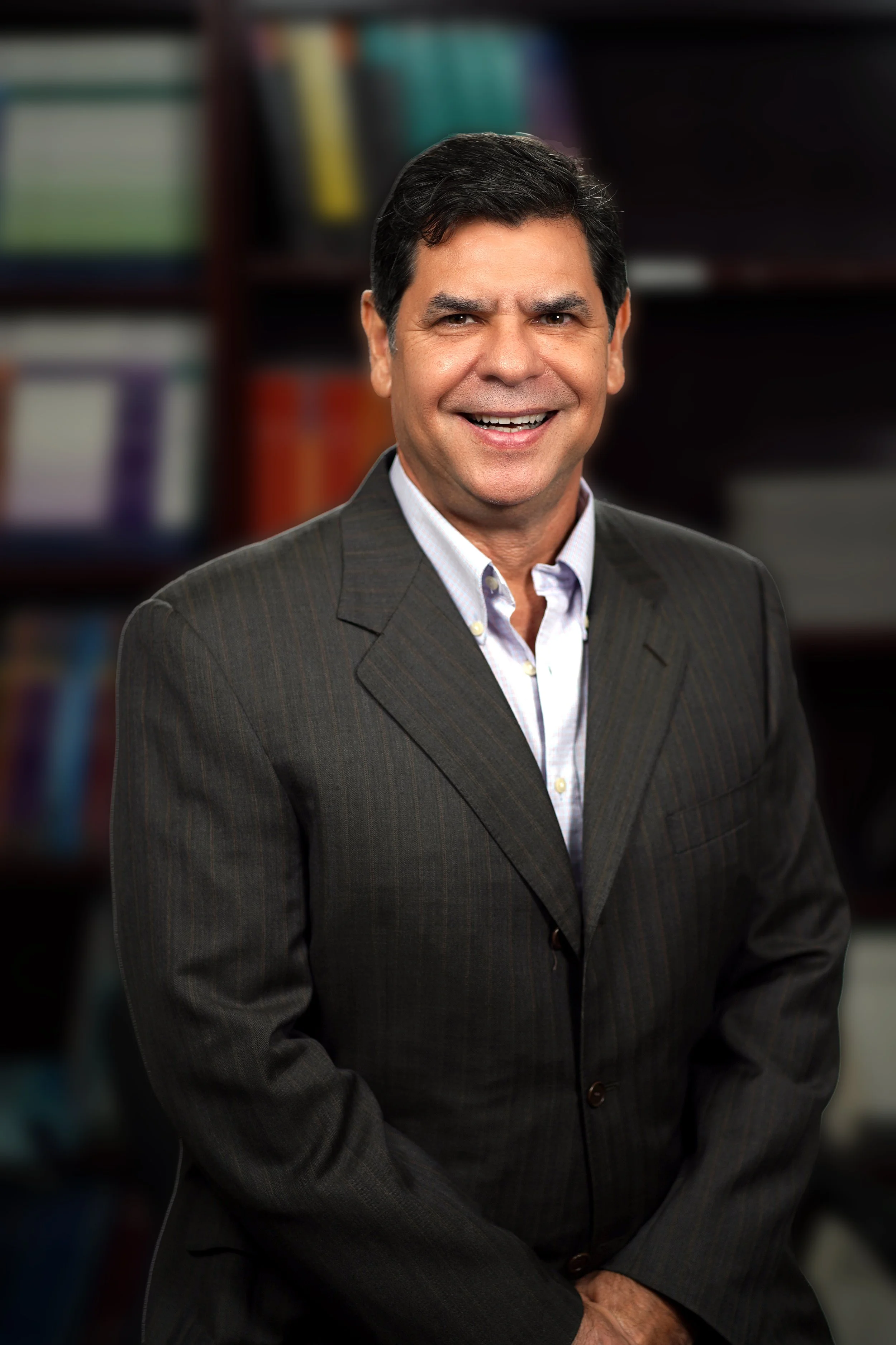 A middle-aged man in a dark suit smiling in a library or office setting with blurred bookshelves in the background.