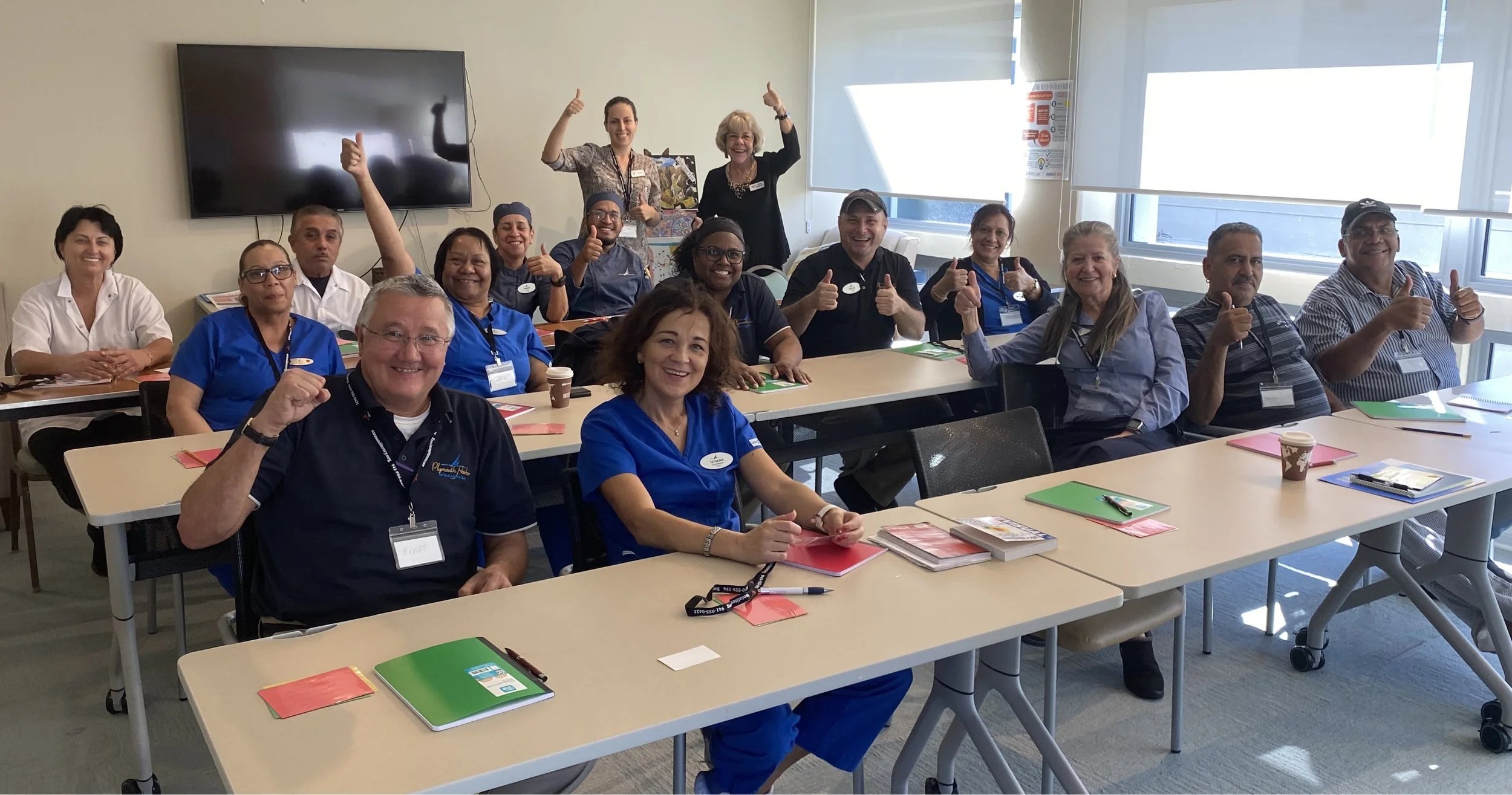 Group photo of diverse healthcare professionals in a meeting room, smiling and giving thumbs up, seated at tables with notebooks and promotional materials.