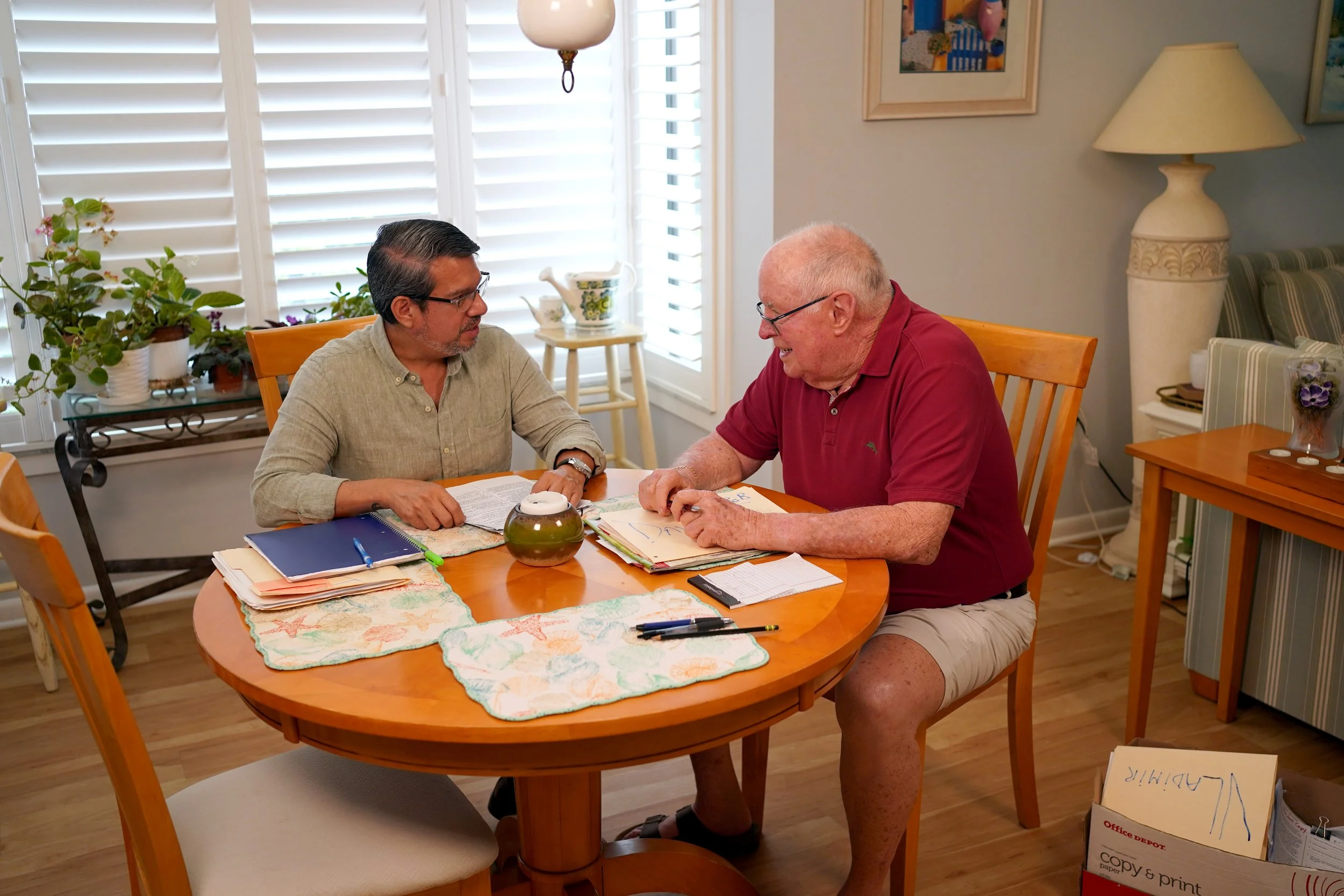 Two men sit at a dining table, engaged in conversation. The man on the left has dark hair, glasses, and is wearing a beige shirt. The man on the right has a shaved head, glasses, and is wearing a red polo shirt. The table has papers, a notebook, and a decorative tablecloth. The background shows a room with white shutters, houseplants, artwork, and a large lamp.