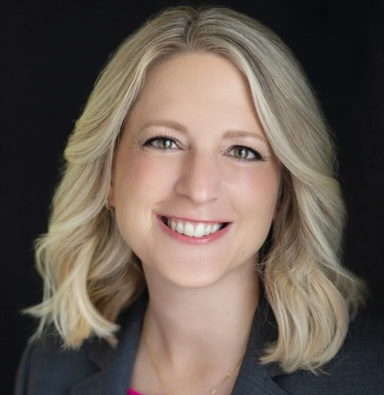 Headshot of a smiling woman with blonde hair, wearing a dark blazer against a black background.