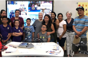 Group of children and adults in a classroom or community center, standing around a table with books and papers, smiling for the camera.