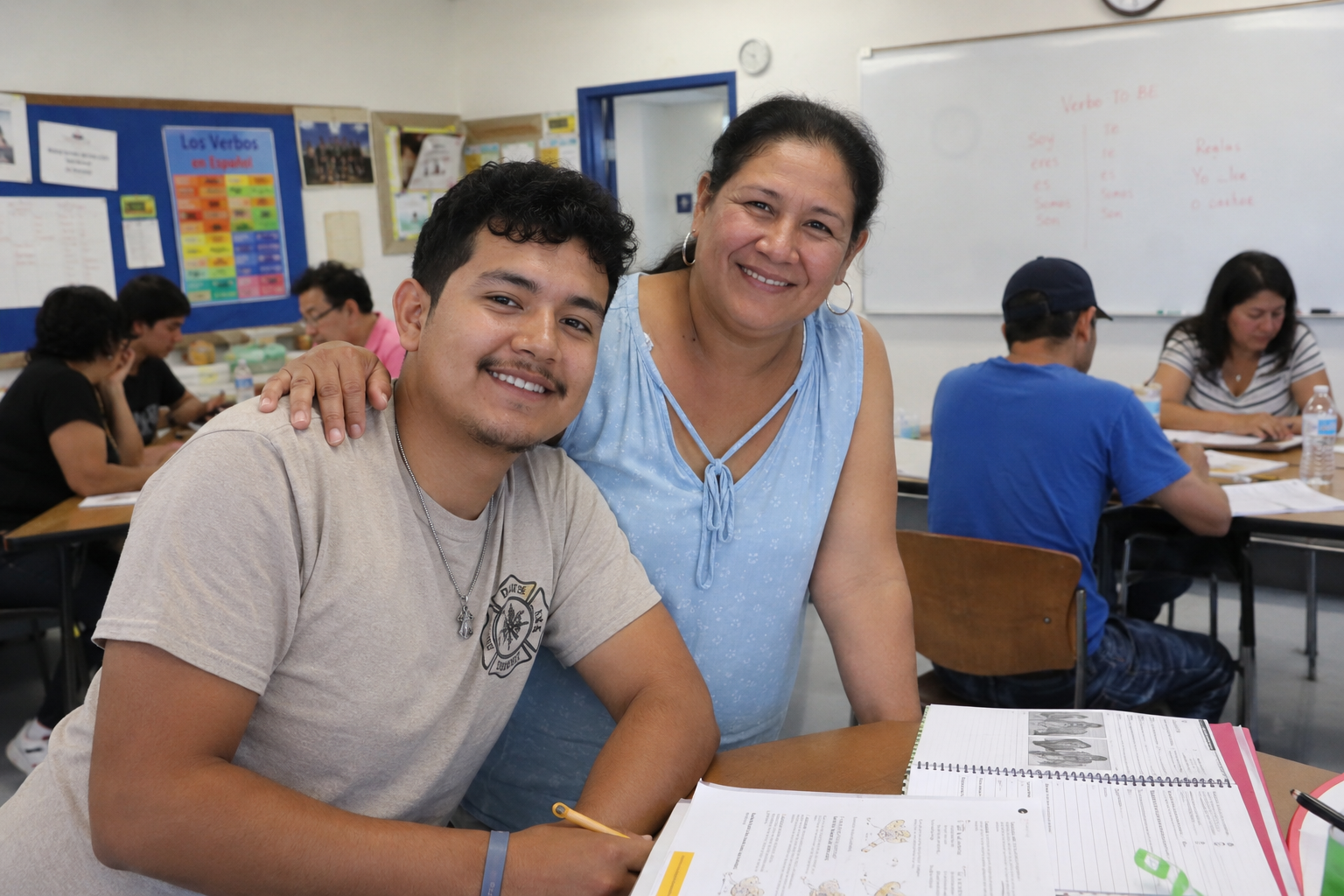 A young man and a woman smiling at the camera in a classroom. The woman has her hand on the young man's shoulder. Other students are sitting at desks in the background, studying or writing.